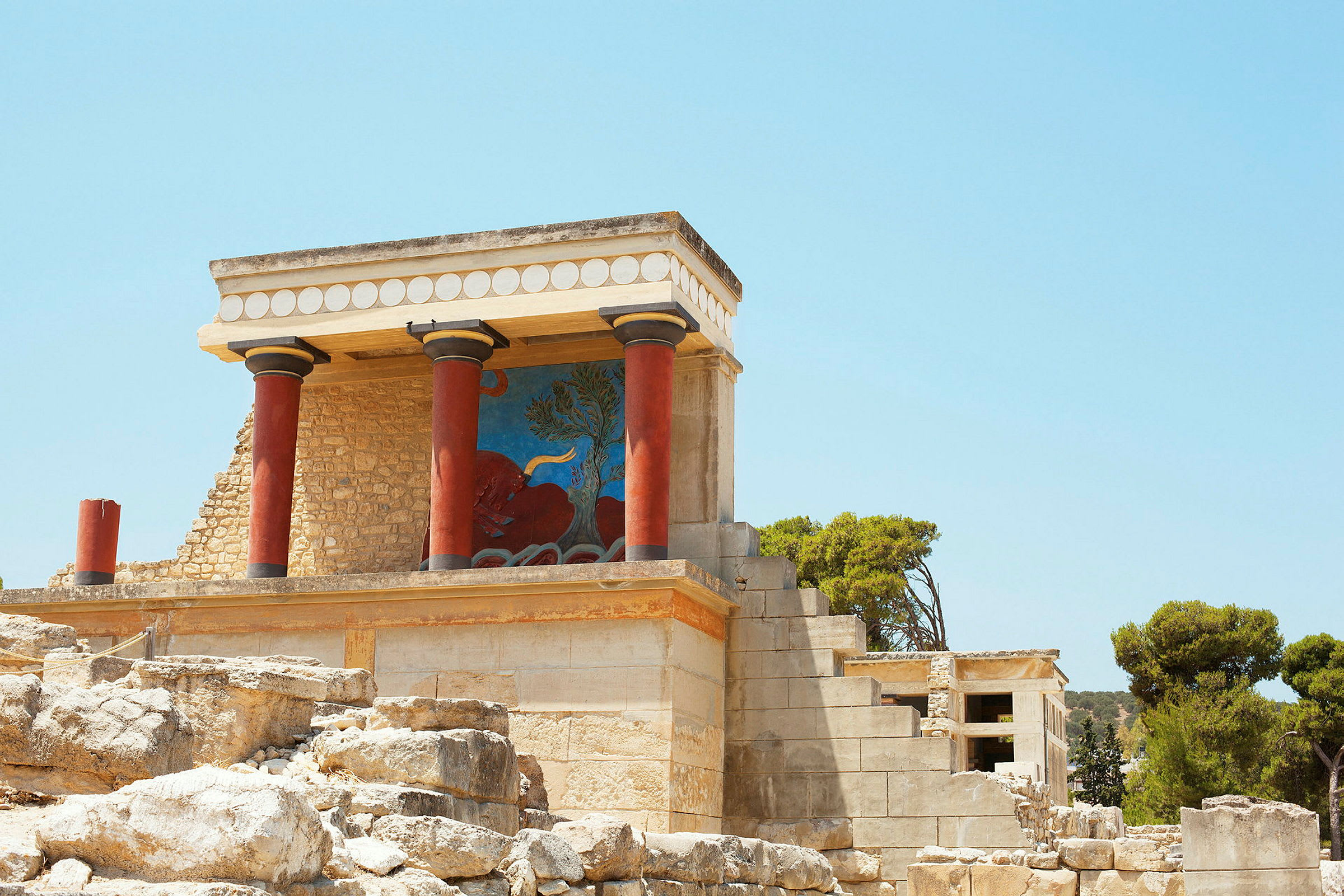 Knossos-palasset med røde søyler og fargerike utsmykninger under en klar blå himmel. (AI-generert tekst)