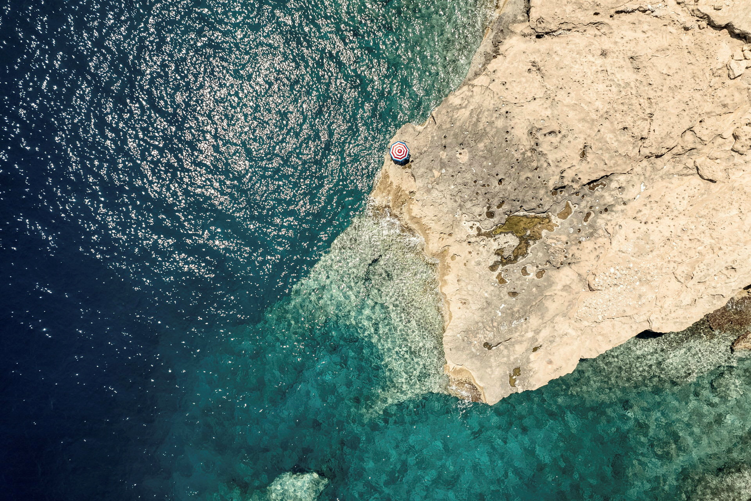 Utsikt ovenfra av en klippe ved havet med en hvit og rød parasoll nær kanten. (AI-generert tekst)