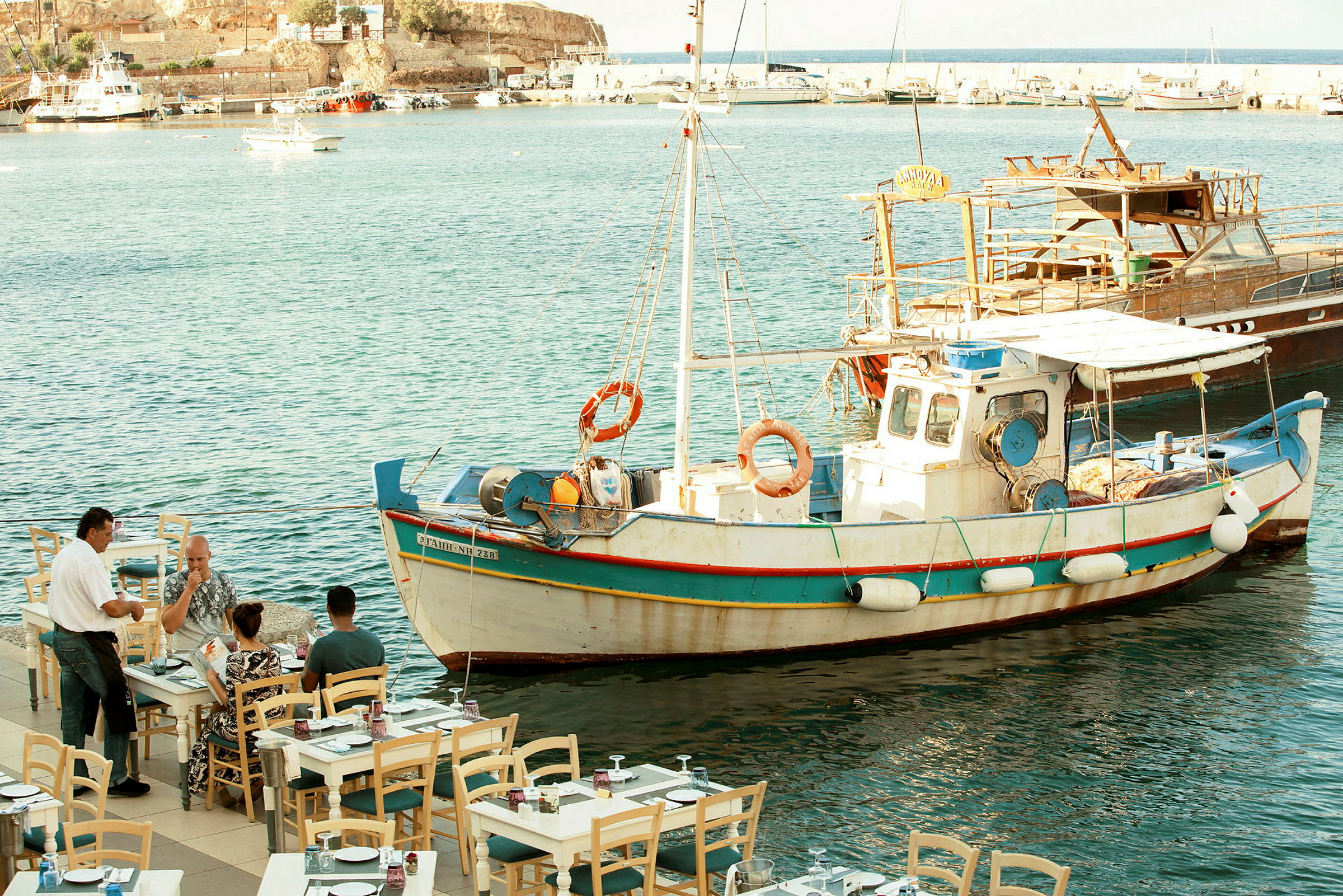 En fiskebåt ligger fortøyd ved en brygge nær en restaurant ved havet. (AI-generert tekst)