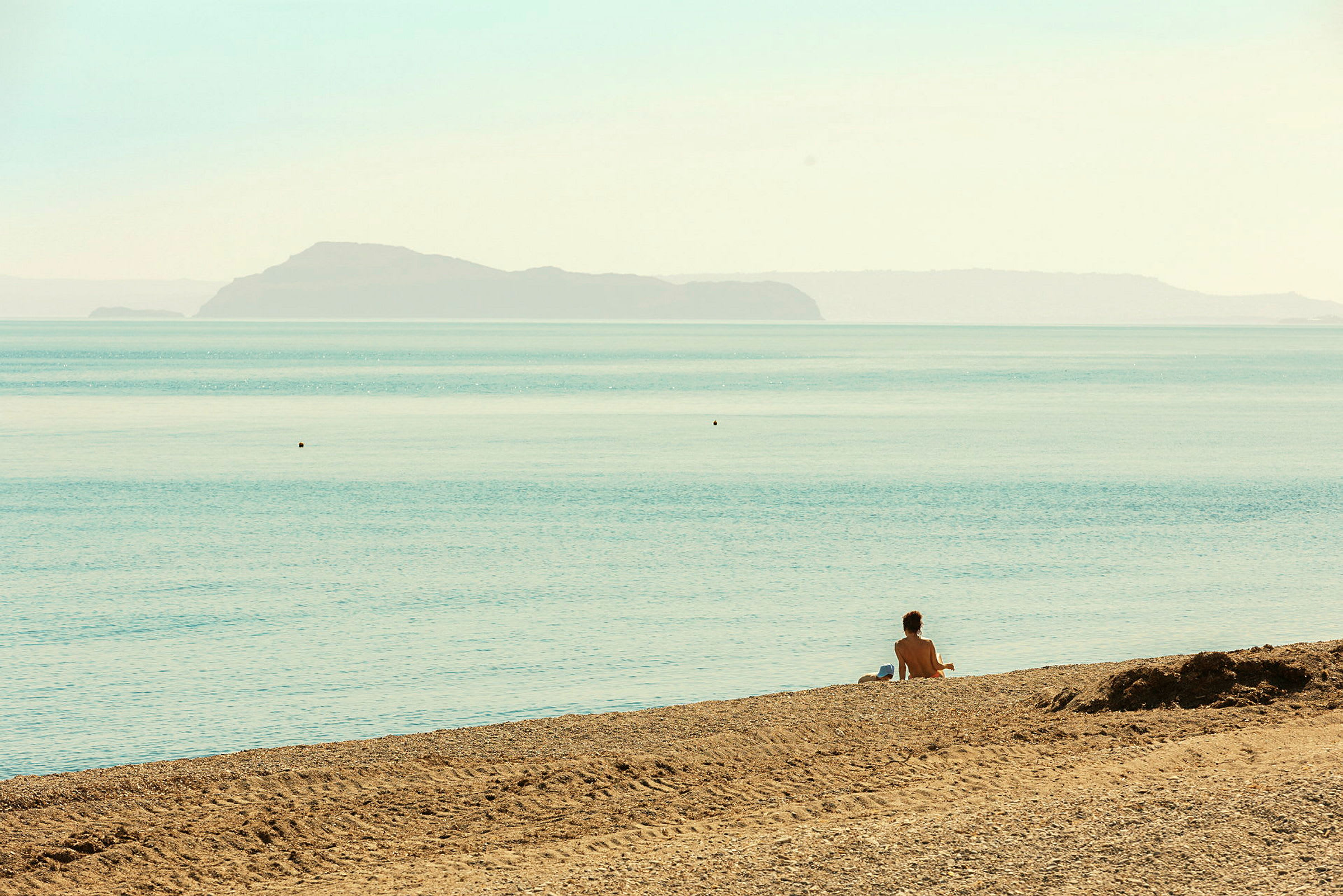 En person sitter på en sandstrand ved havet med en øy i bakgrunnen. (AI-generert tekst)