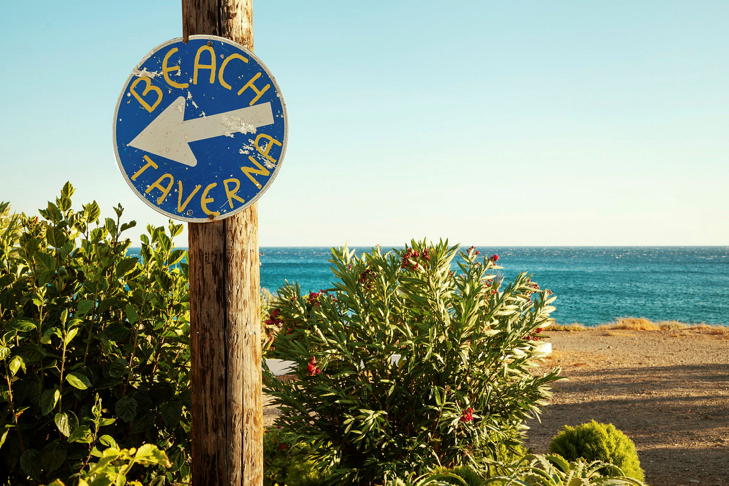 Et skilt med teksten 'Beach Taverna' peker mot stranden, med vegetasjon og havet i bakgrunnen. (AI-generert tekst)