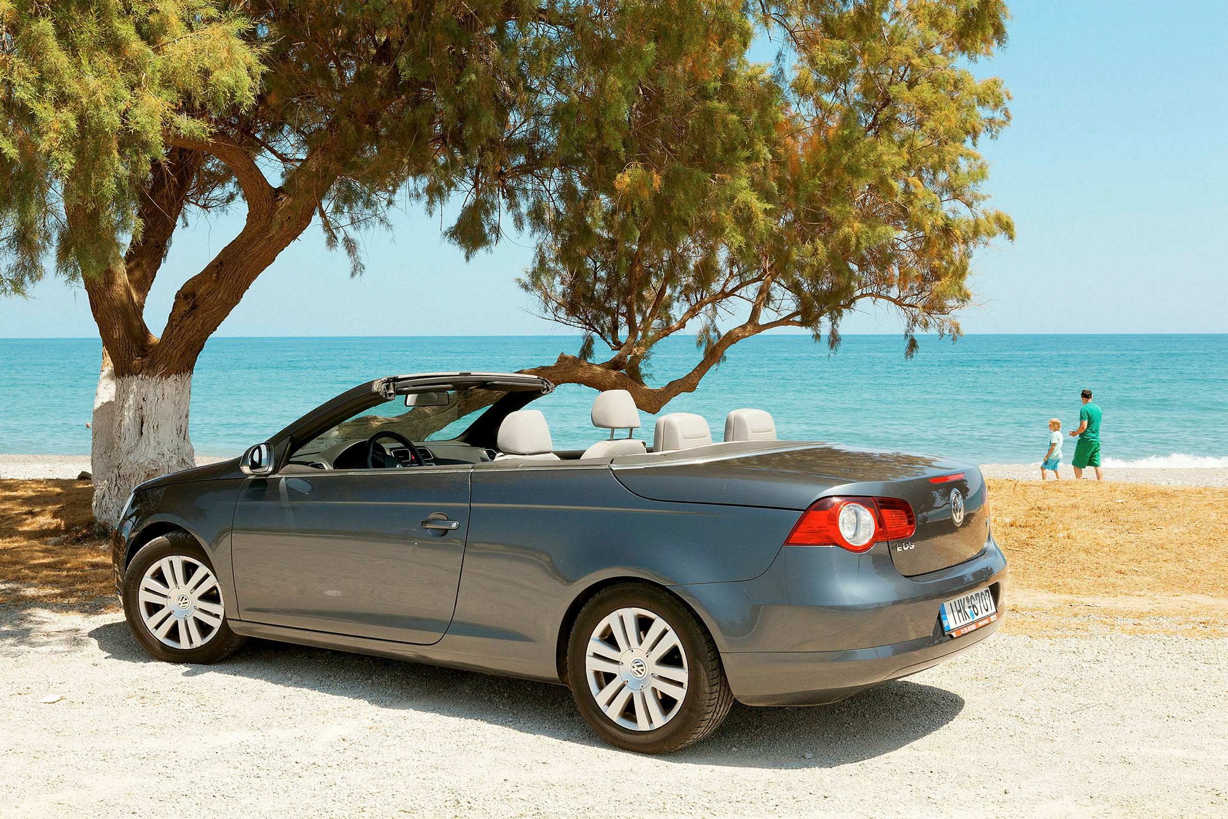 En grå cabriolet er parkert ved en strand med hav og trær i bakgrunnen. (AI-generert tekst)