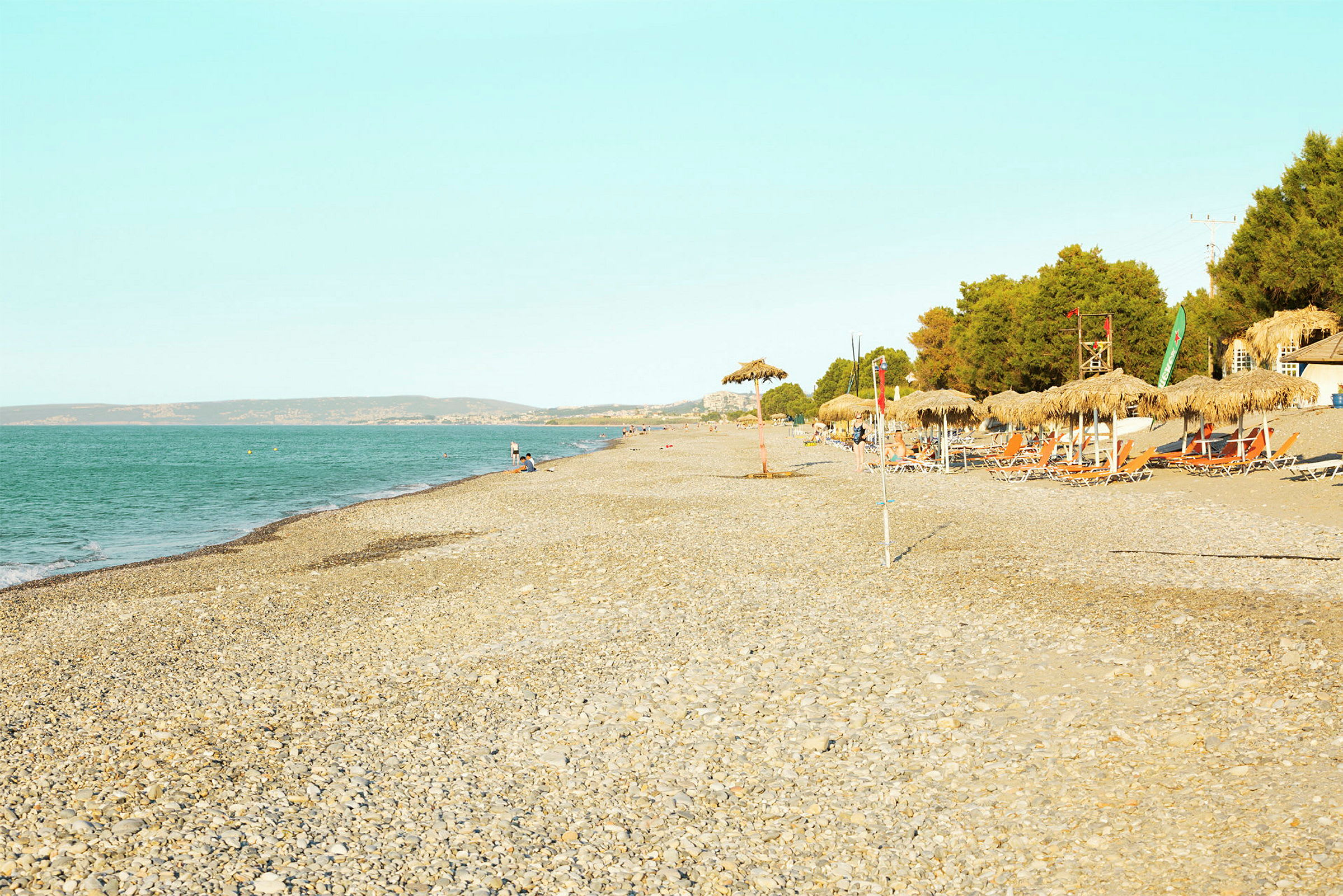 En strand med stein, parasoller og solsenger ved siden av en skog. Havet er synlig til venstre. (AI-generert tekst)