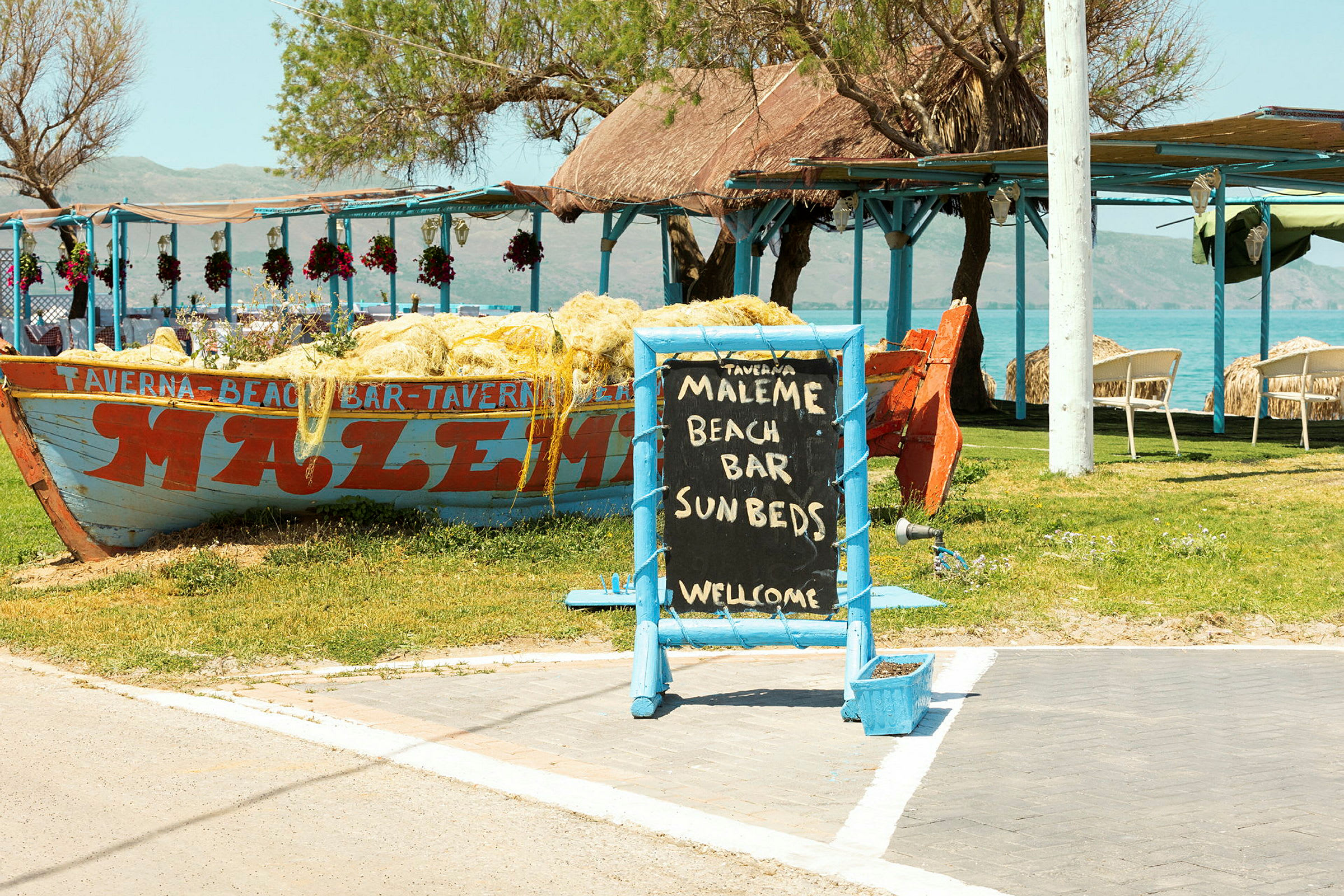 Skilt med teksten 'Maleme Beach Bar Sunbeds Welcome' foran en båt dekorert med nett og en utendørs servering ved havet. (AI-generert tekst)