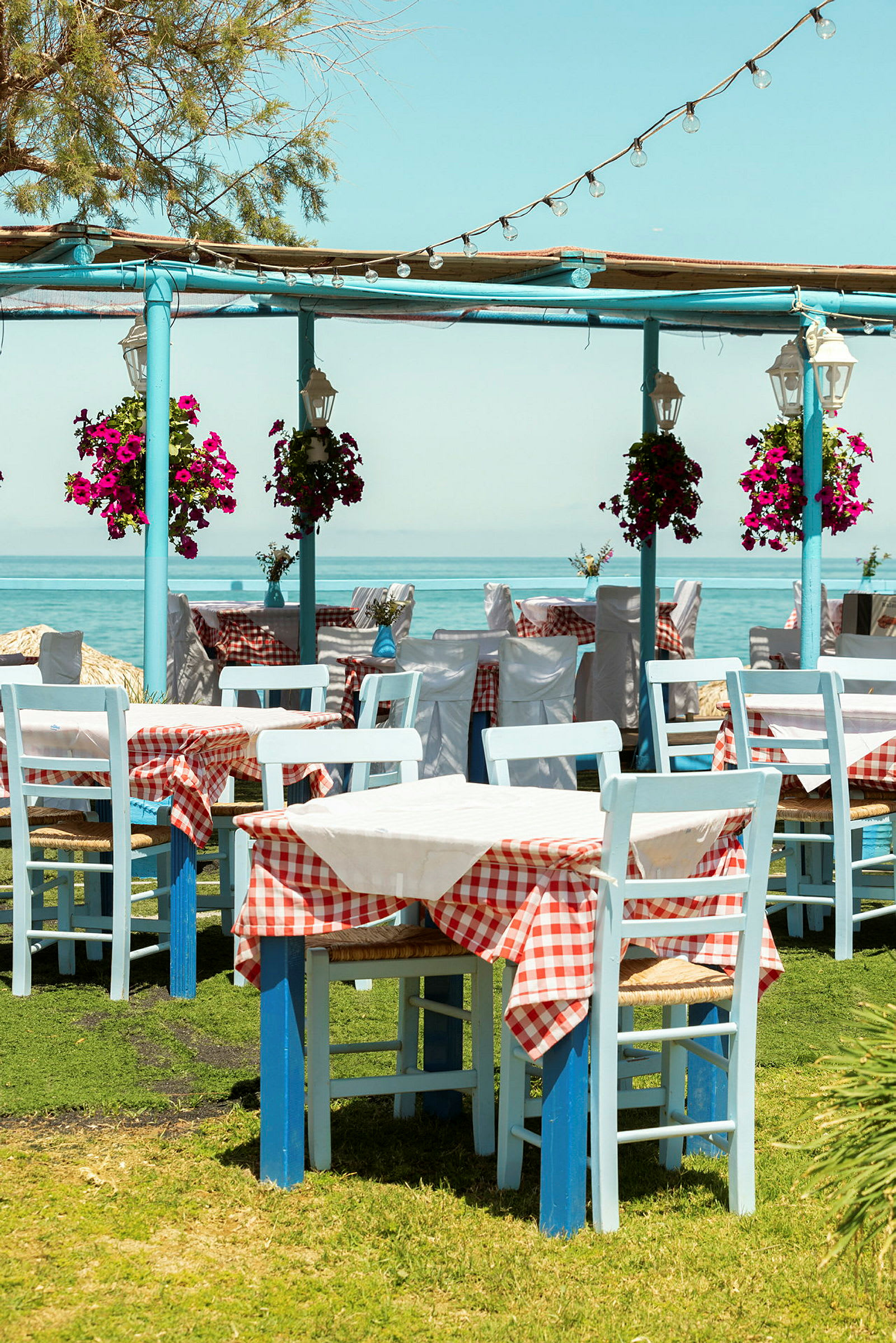 Bilde av tomme restaurantbord med hvite og rødrutete duker, blå stoler og blomsterpotter på gress nær havet. (AI-generert tekst)