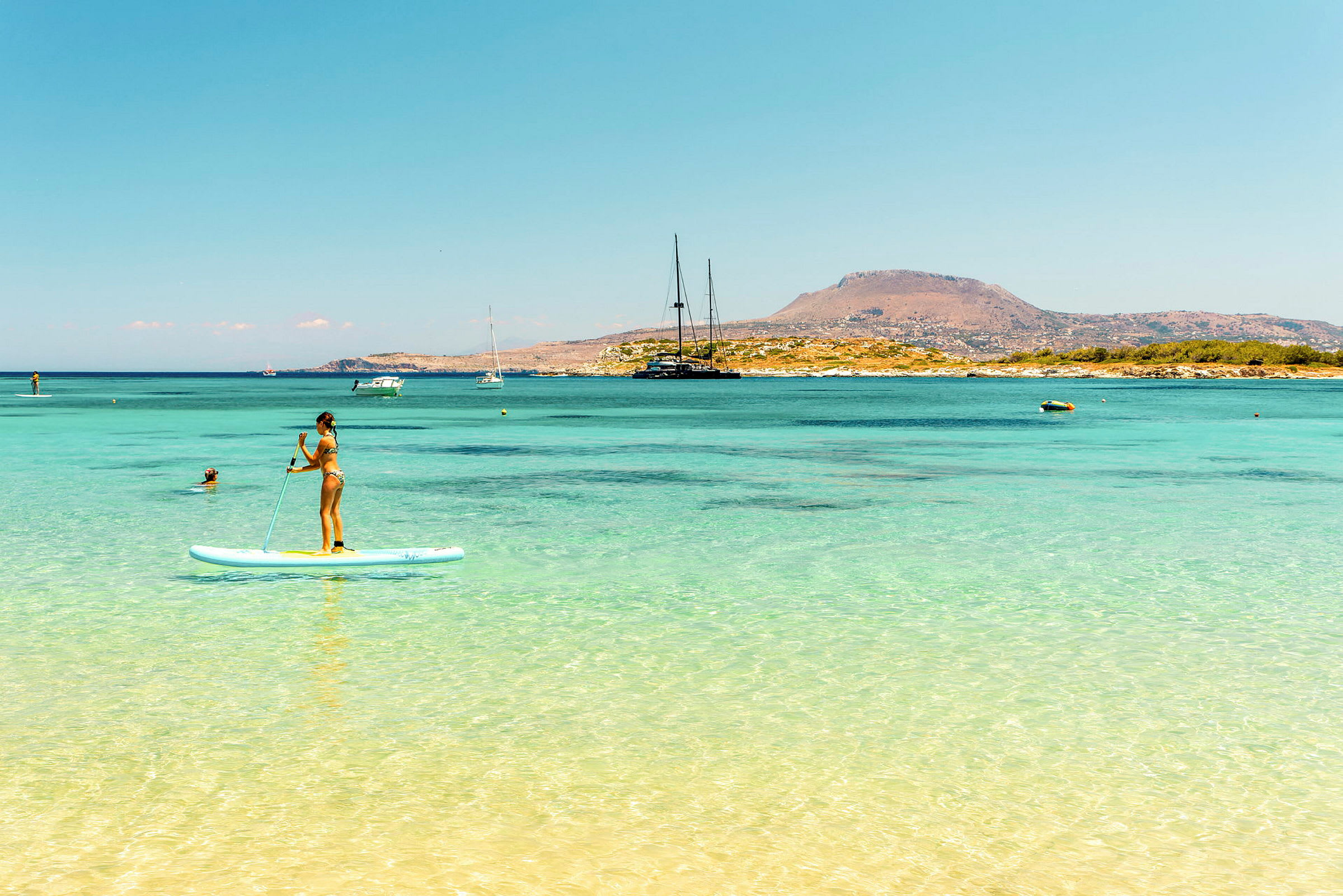 Person som står på et paddleboard i klart blått vann med en fjellrik kystlinje i bakgrunnen. (AI-generert tekst)