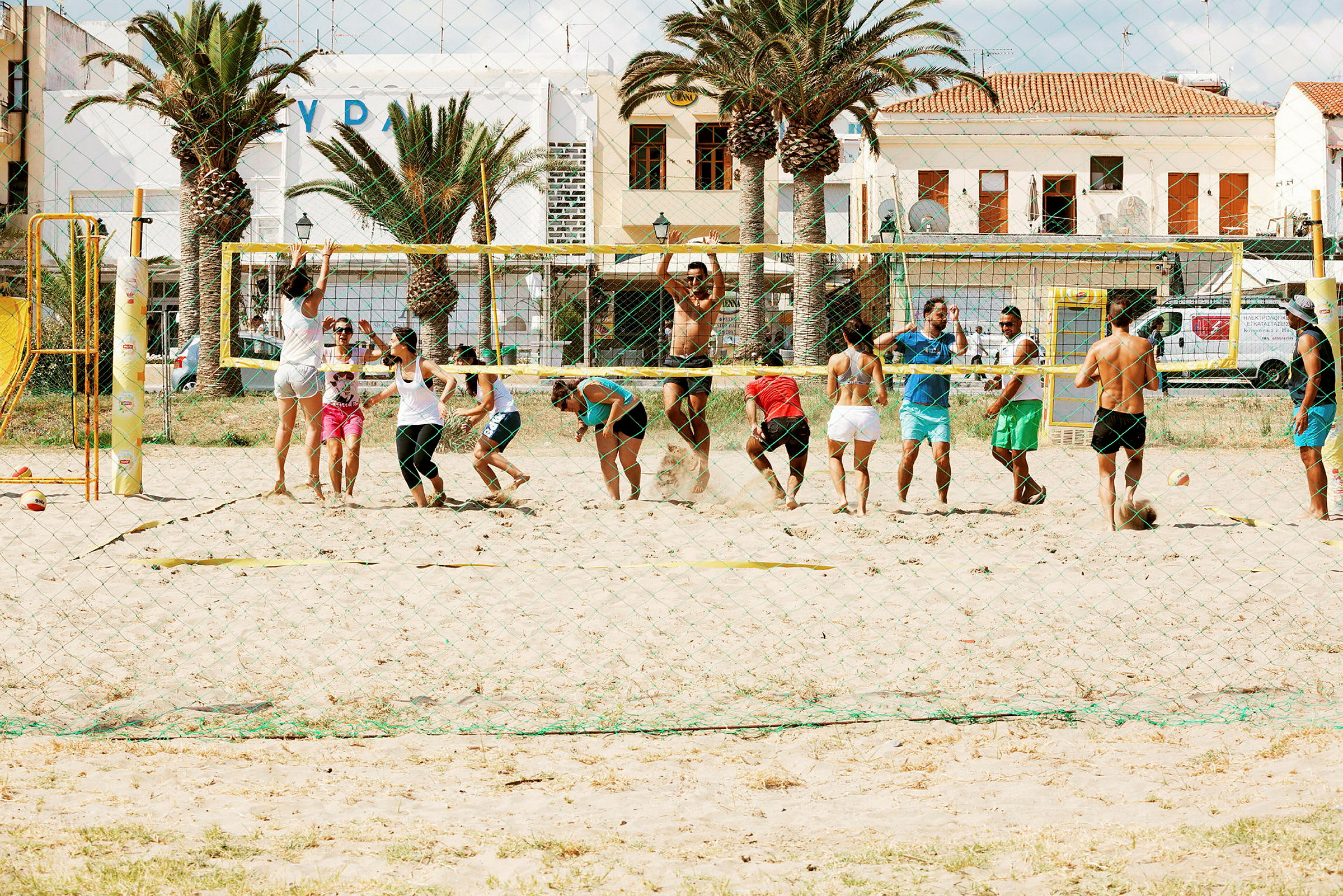 En gruppe mennesker spiller strandvolleyball på en sandbane foran bygninger og palmer. (AI-generert tekst)