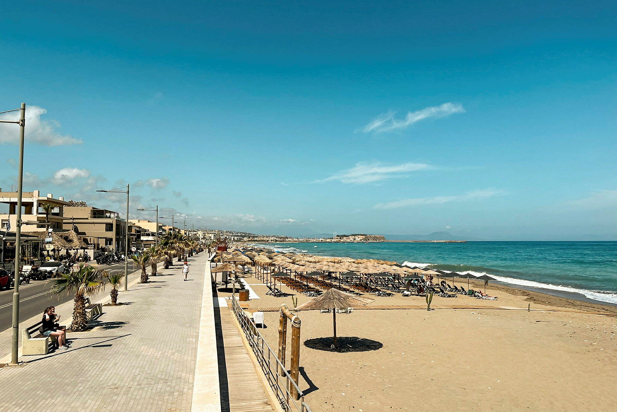 En strand med parasoller og solsenger ved siden av en strandpromenade og små bygninger. Havet er synlig i bakgrunnen. (AI-generert tekst)