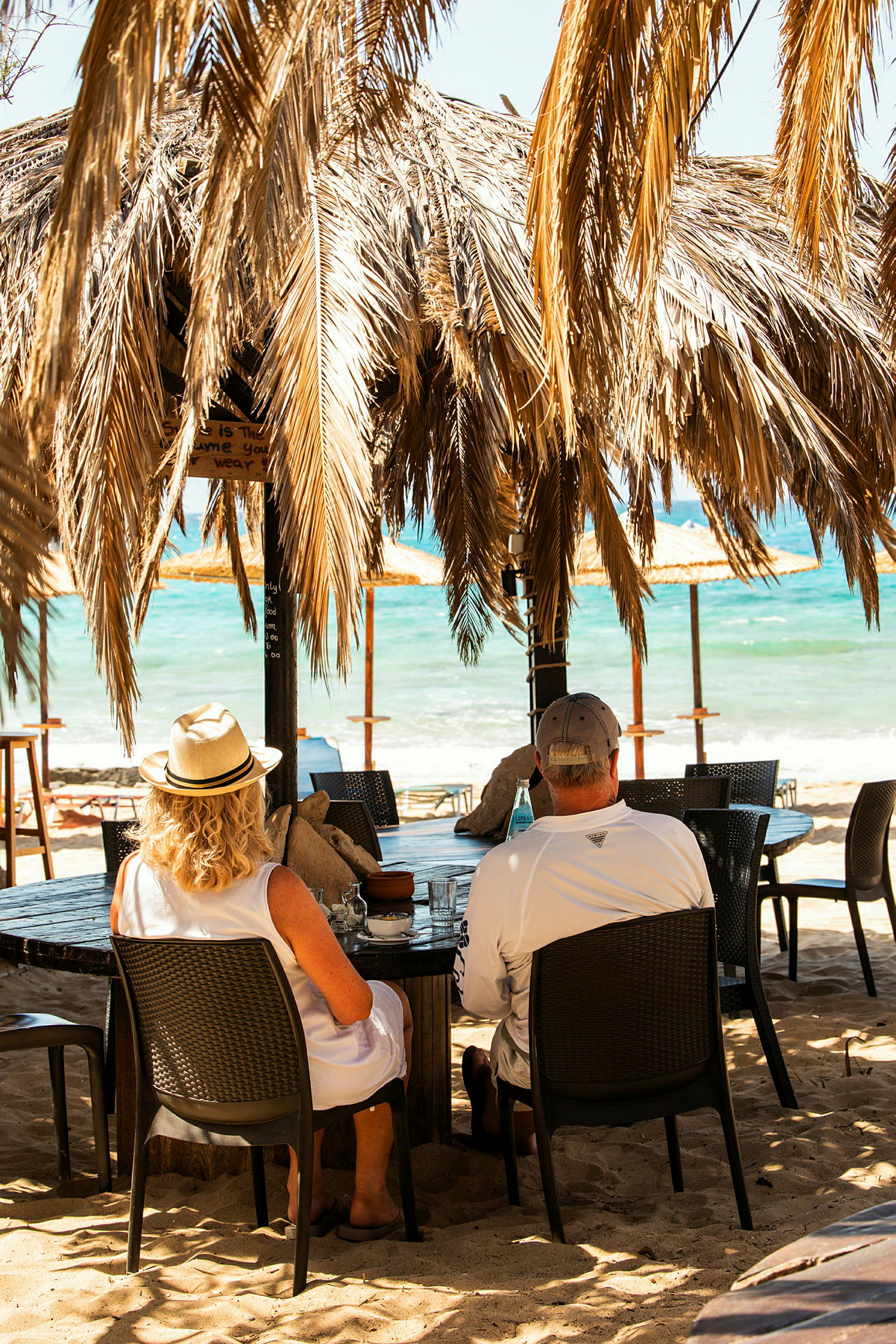 To personer sitter ved et bord under en takstruktur med palmeblad, med utsikt over en strand og havet. (AI-generert tekst)