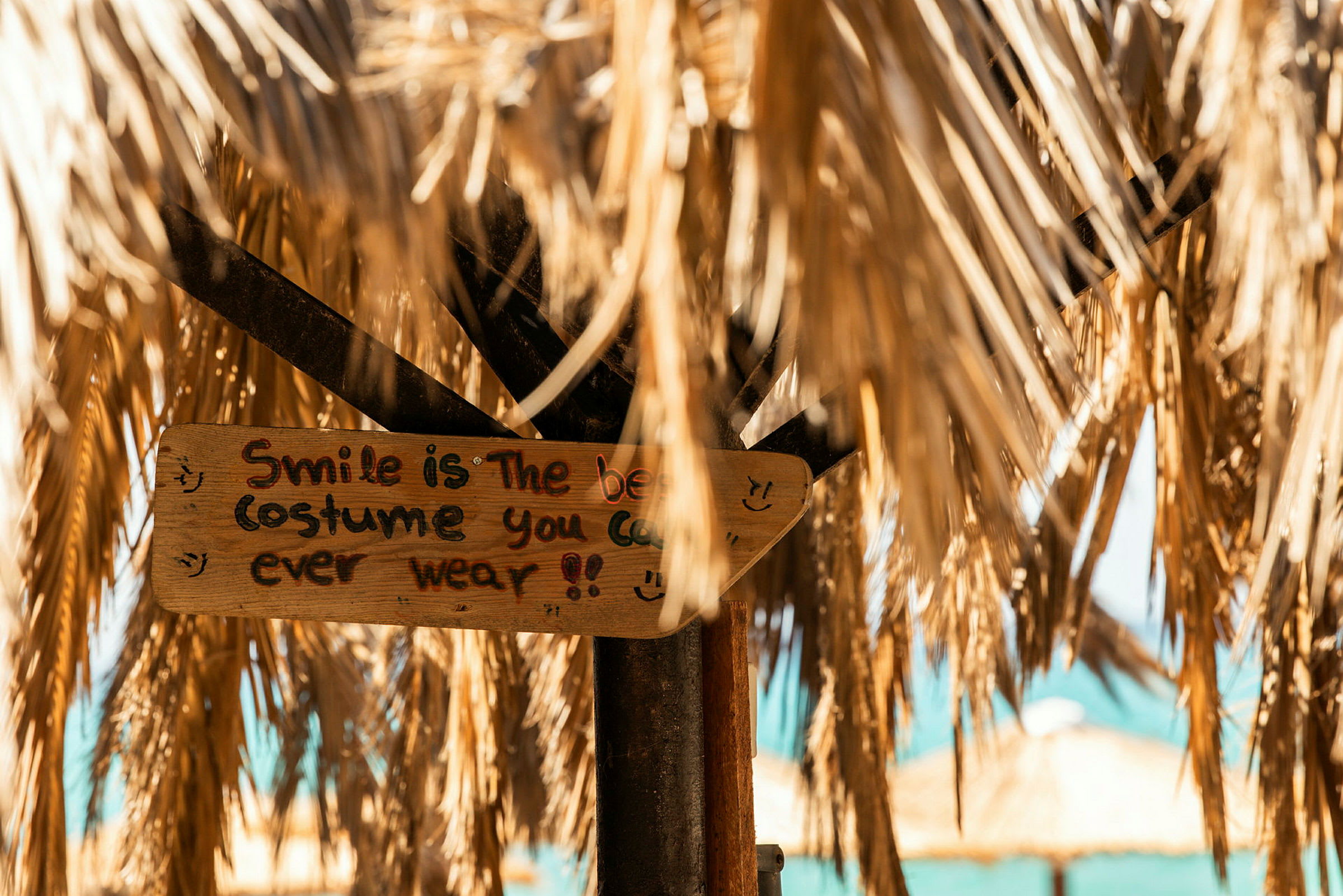 Et nærbilde av et håndmalt treskilt som henger under tørkede palmebladtak. Skiltet har teksten "Smile is the best costume you can ever wear!". (AI-generert tekst)