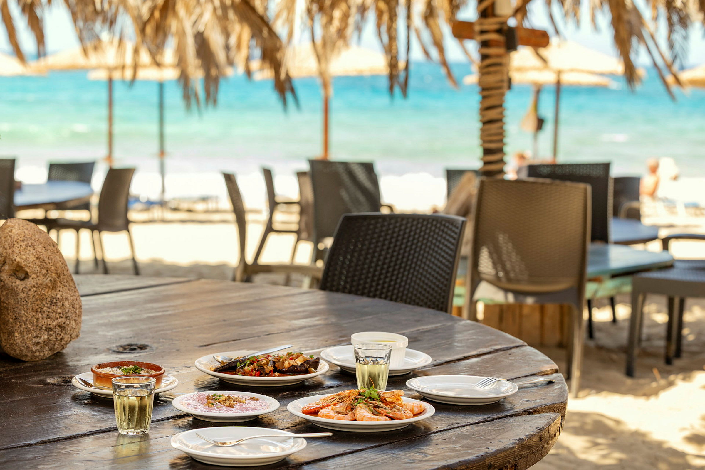 Et bord på en strandrestaurant med flere tallerkener med mat og drikke. Bakgrunnen viser sandstrand og havet. (AI-generert tekst)
