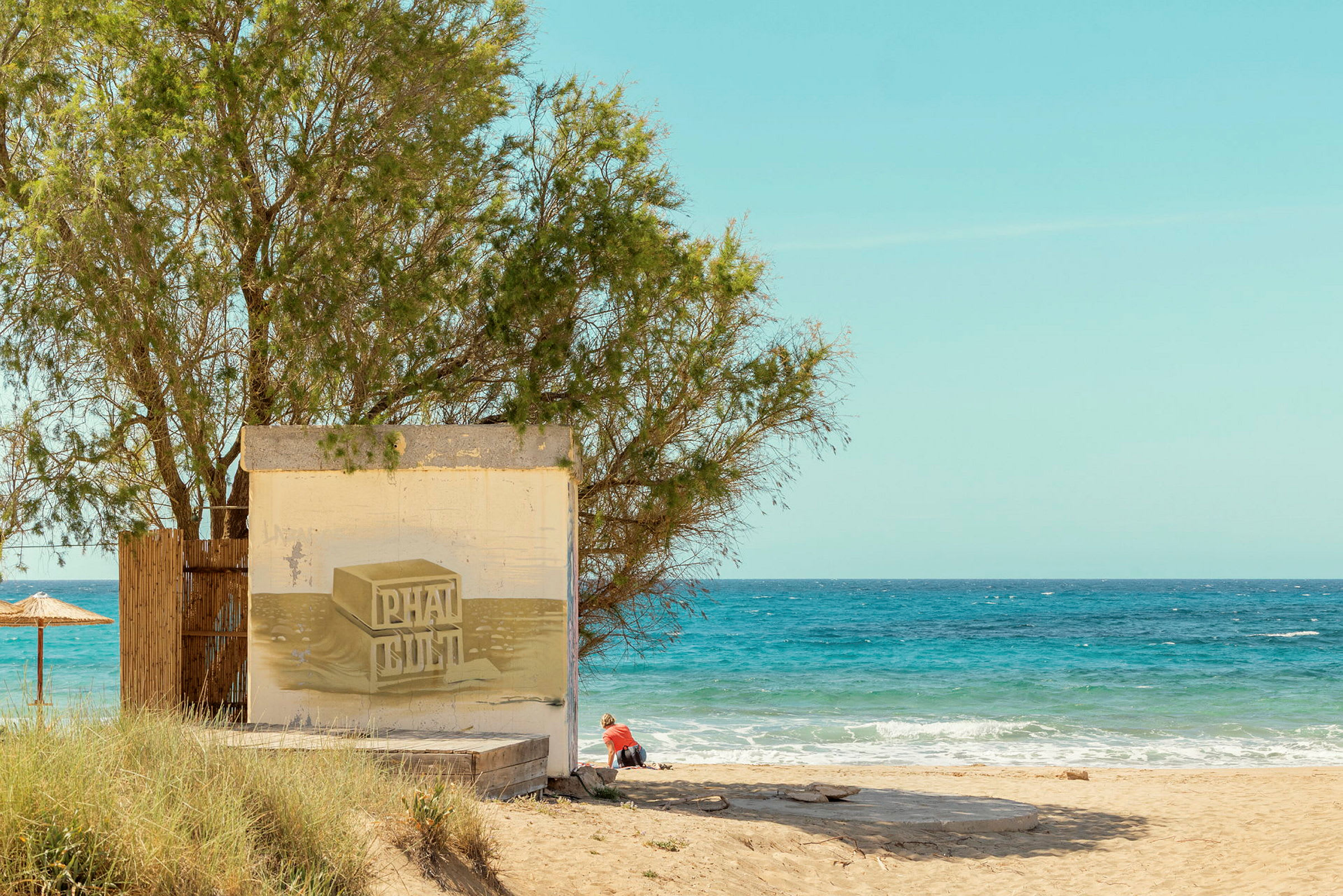 En strand med en lav bygning til venstre dekorert med kunstverk. Et tre står ved siden av bygningen, og en person sitter i sanden. Havet er synlig i bakgrunnen. (AI-generert tekst)