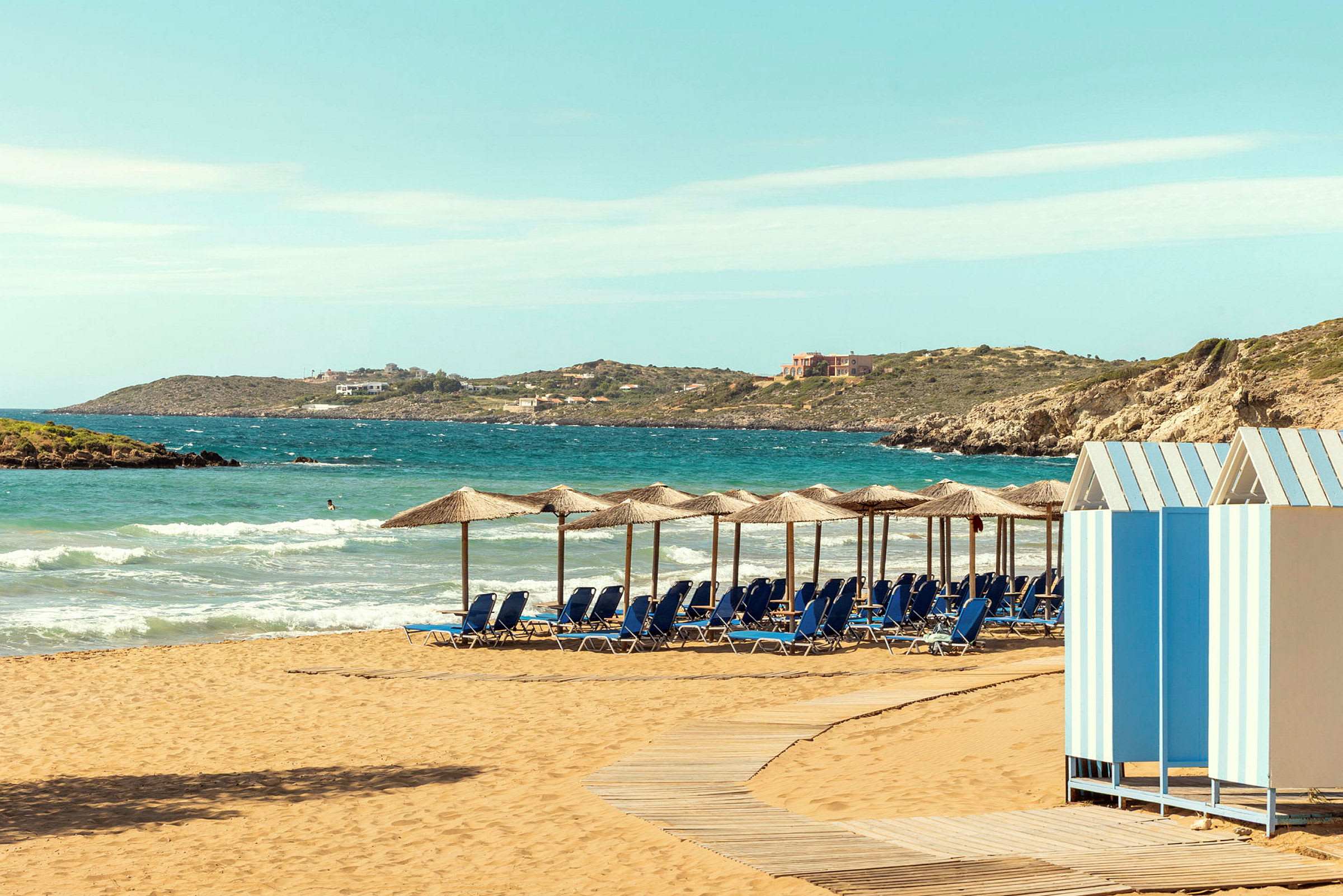 Strand med parasoller og solsenger, en trepromenade og små fargerike strandhytter. (AI-generert tekst)