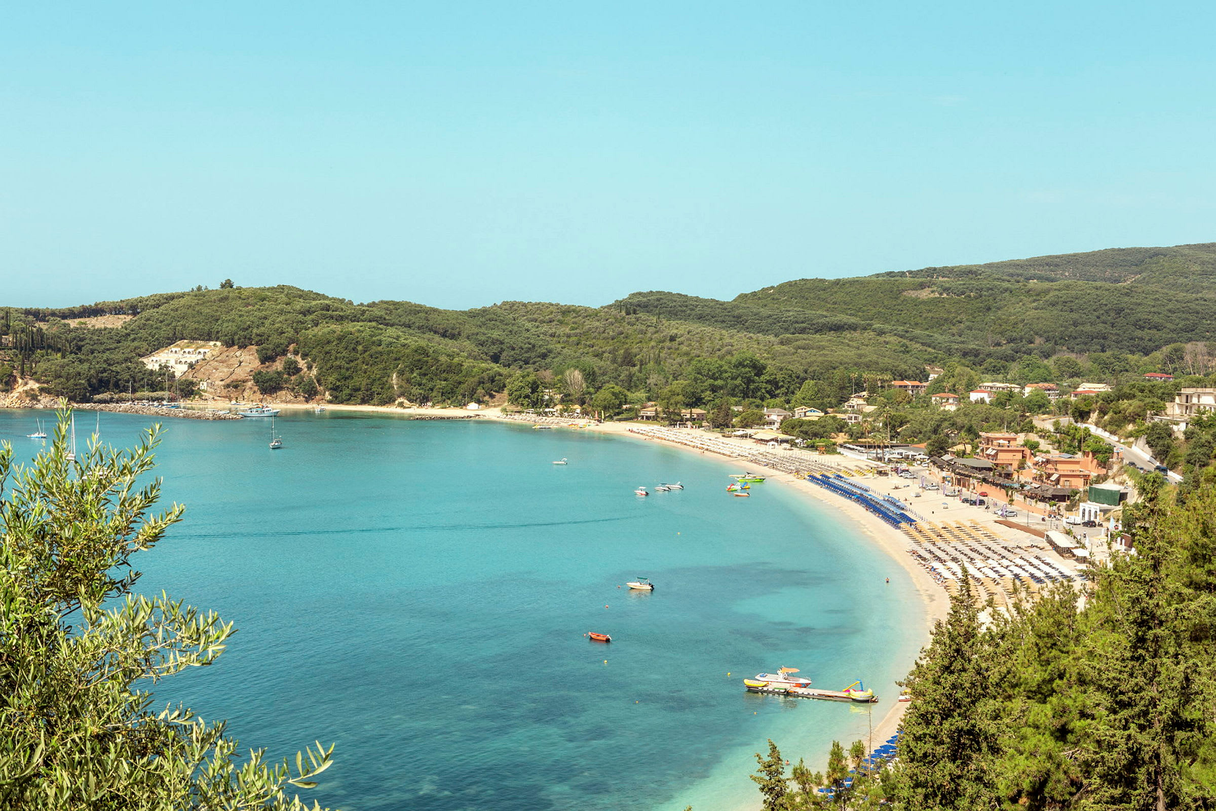 En bukt med sandstrand, solsenger og turkist vann, og skogkledd landskap i bakgrunnen. (AI-generert tekst)