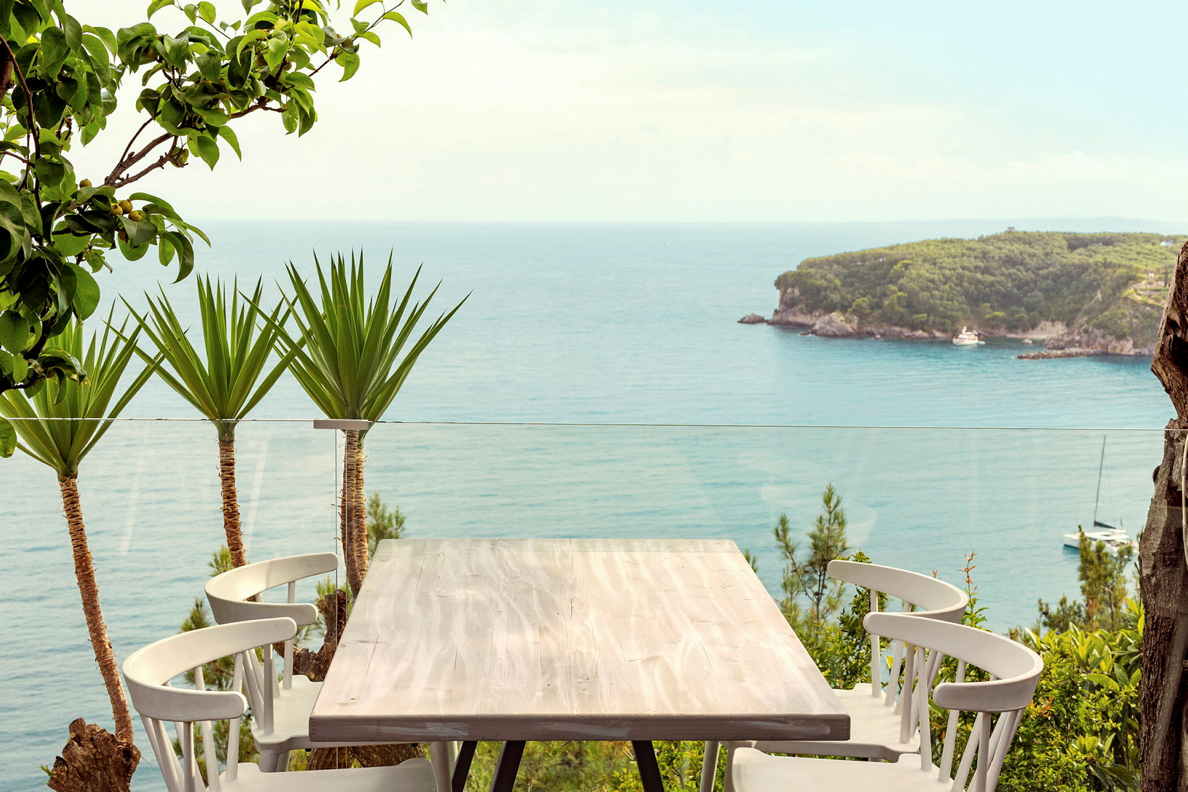 Bord med stoler på en terrasse med utsikt over havet og en grønn skogkledd odde. (AI-generert tekst)