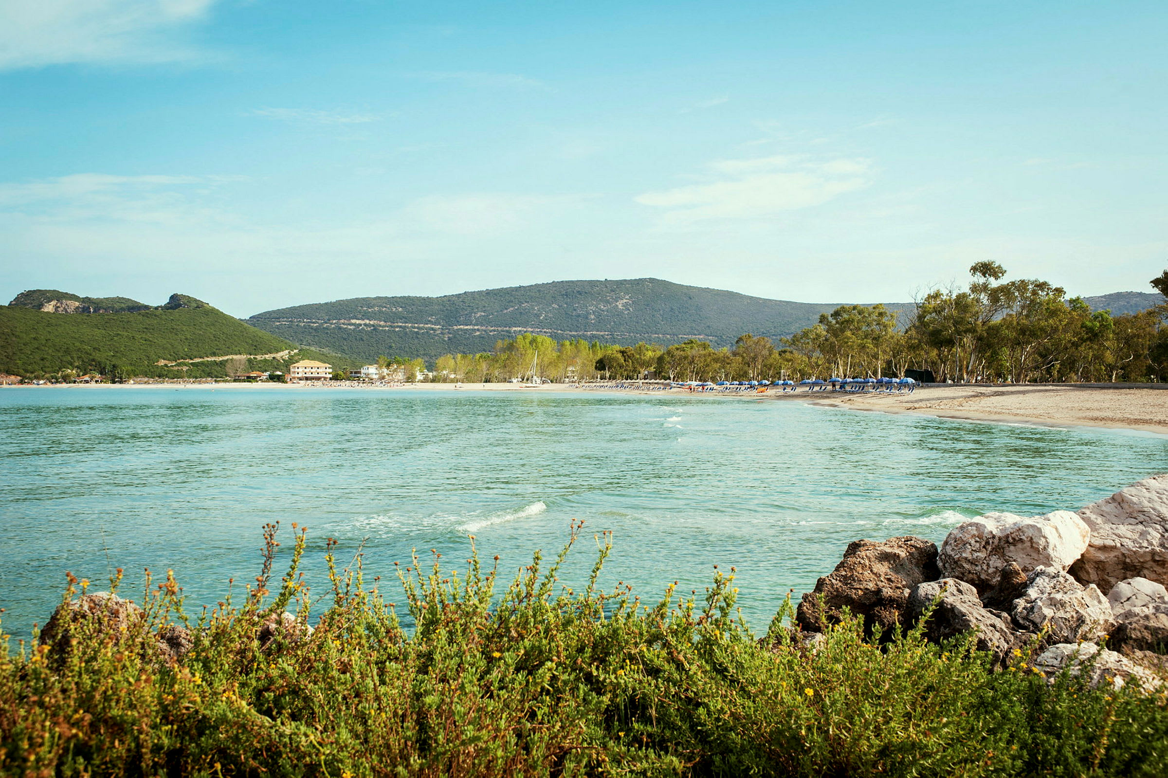 En strand med klart vann, grønne åser og trær i bakgrunnen. (AI-generert tekst)
