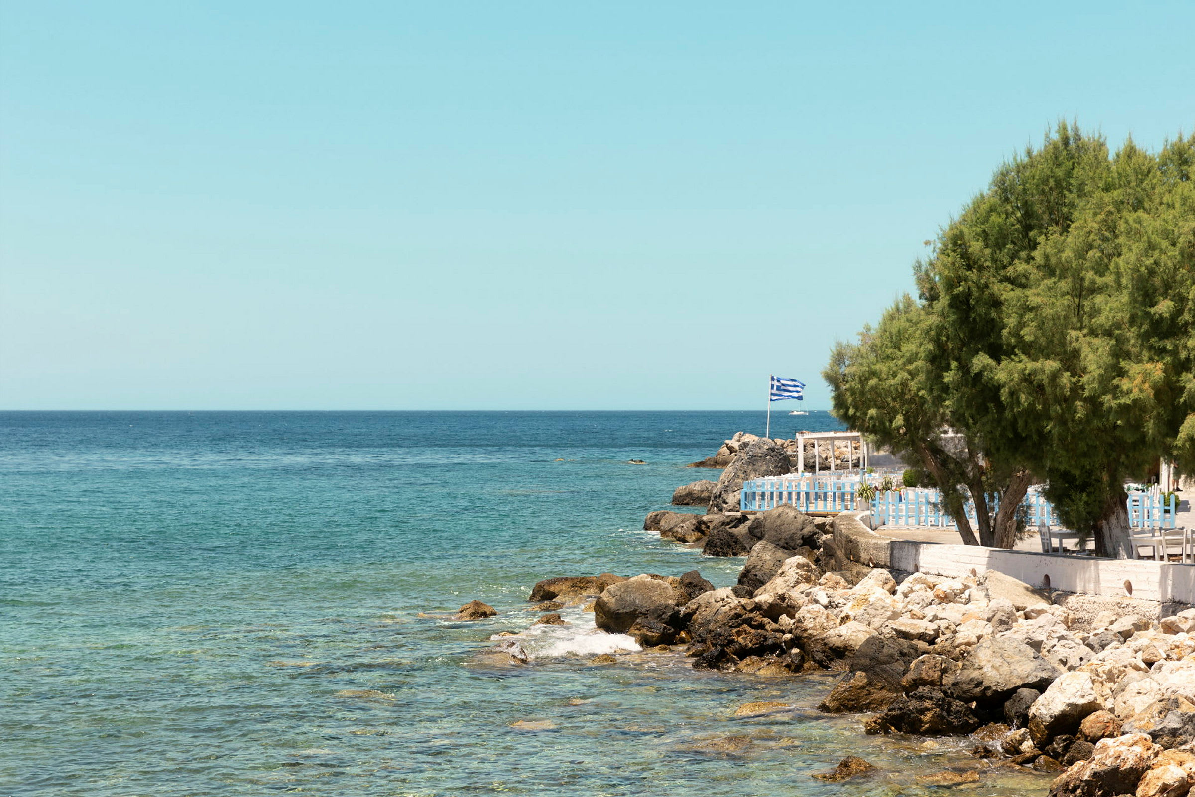 Utsikt over havet med klipper og en bygning med gresk flagg på stranden. (AI-generert tekst)