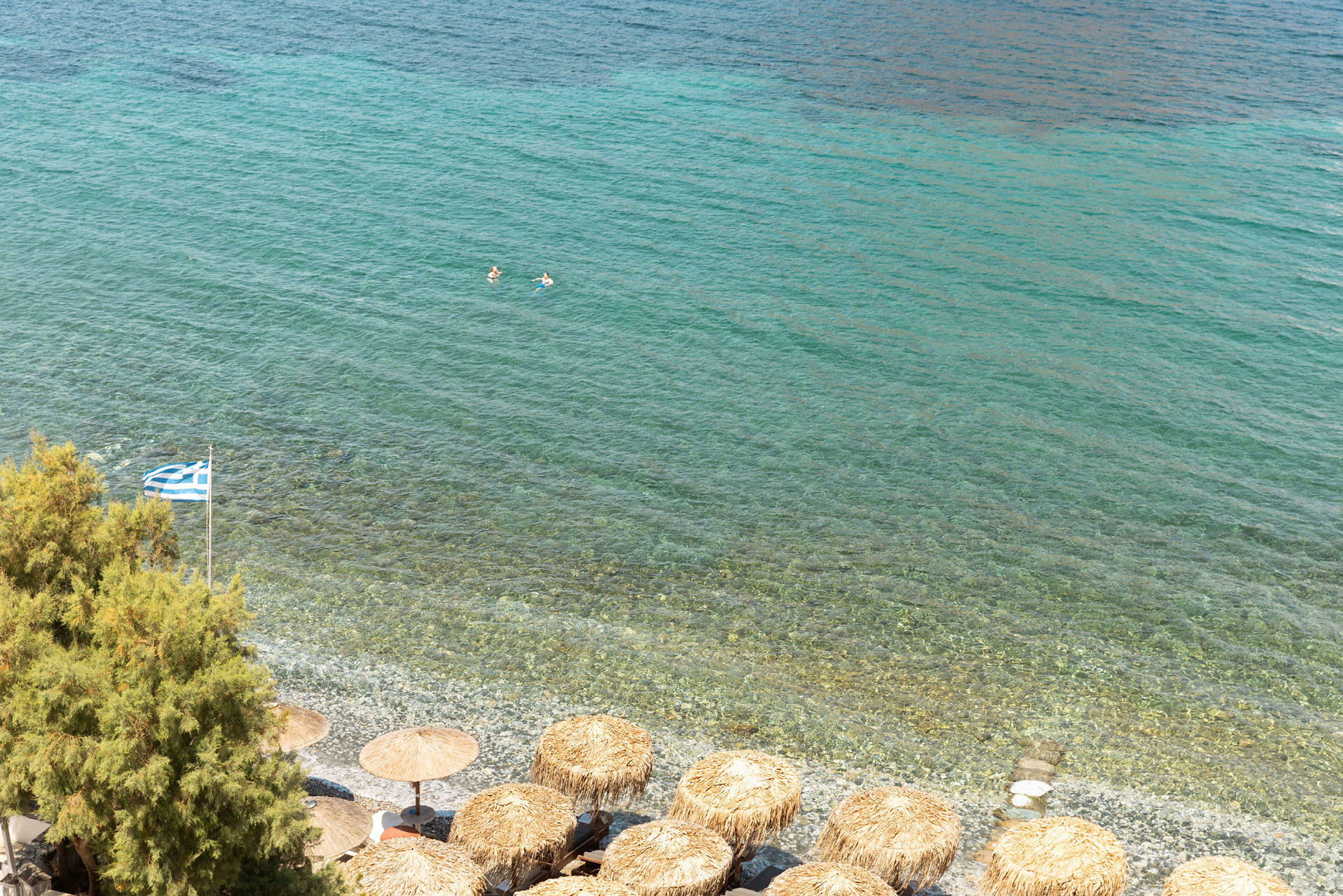 Utsikt over en strand med parasoller, gresk flagg og det klare blå vannet i havet. (AI-generert tekst)