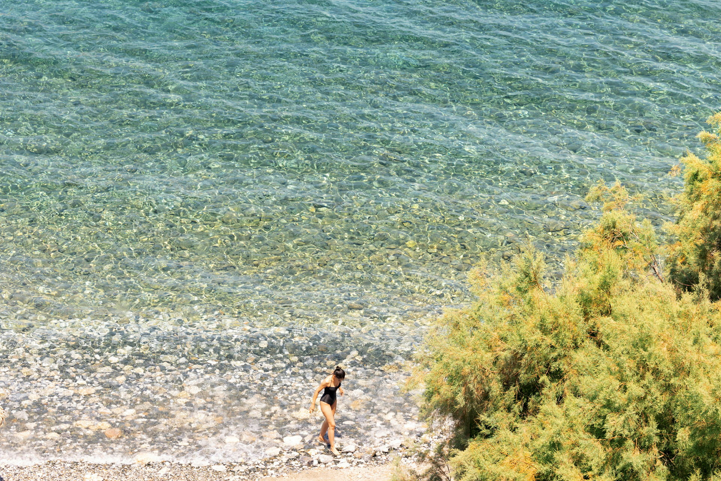 Person i svart badedrakt går på en steinstrand ved klart vann. (AI-generert tekst)
