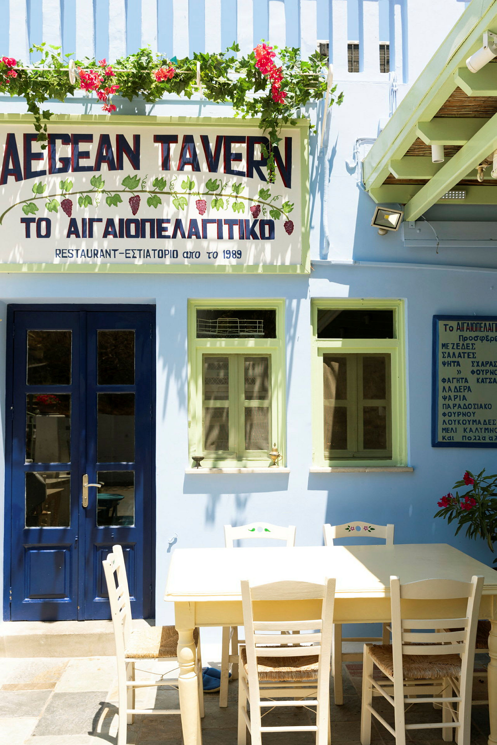 En bygning med blå dør og grønne vinduer, et skilt med teksten 'AEGEAN TAVERN' og et bord med stoler foran. (AI-generert tekst)