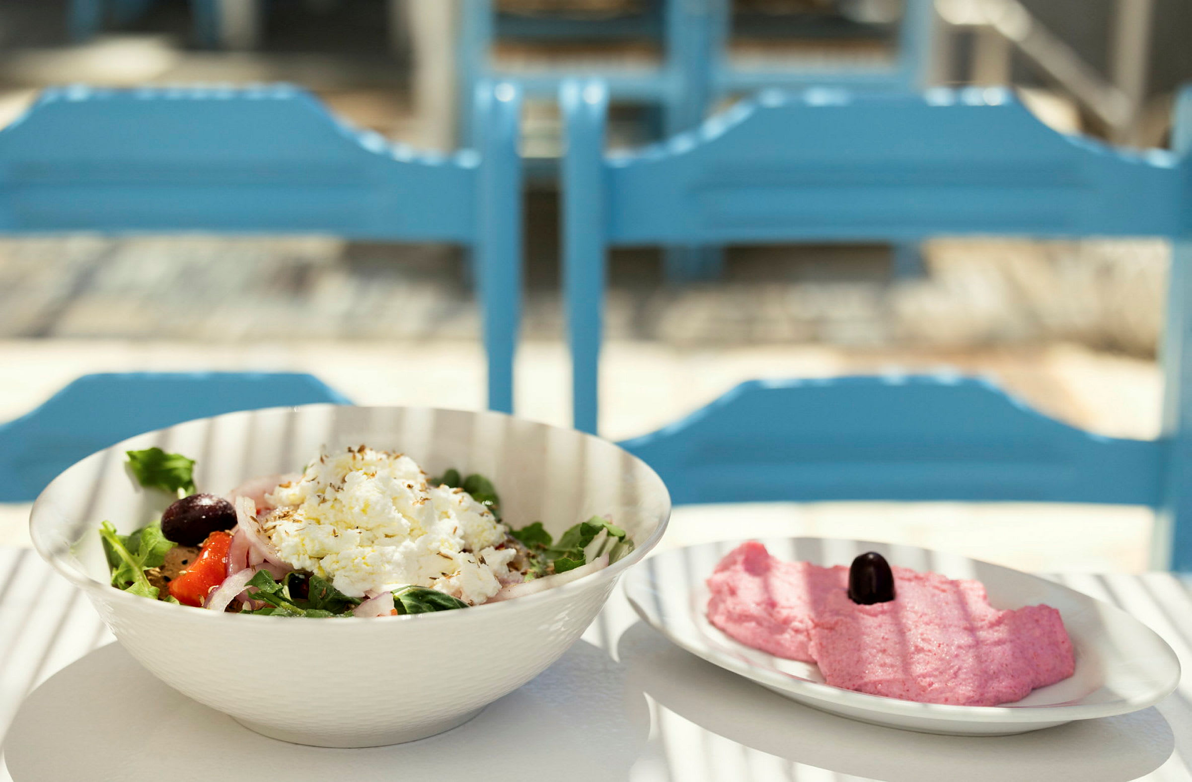 En bolle med salat og en tallerken med rosa røre samt en oliven på et bord med blå stoler i bakgrunnen. (AI-generert tekst)