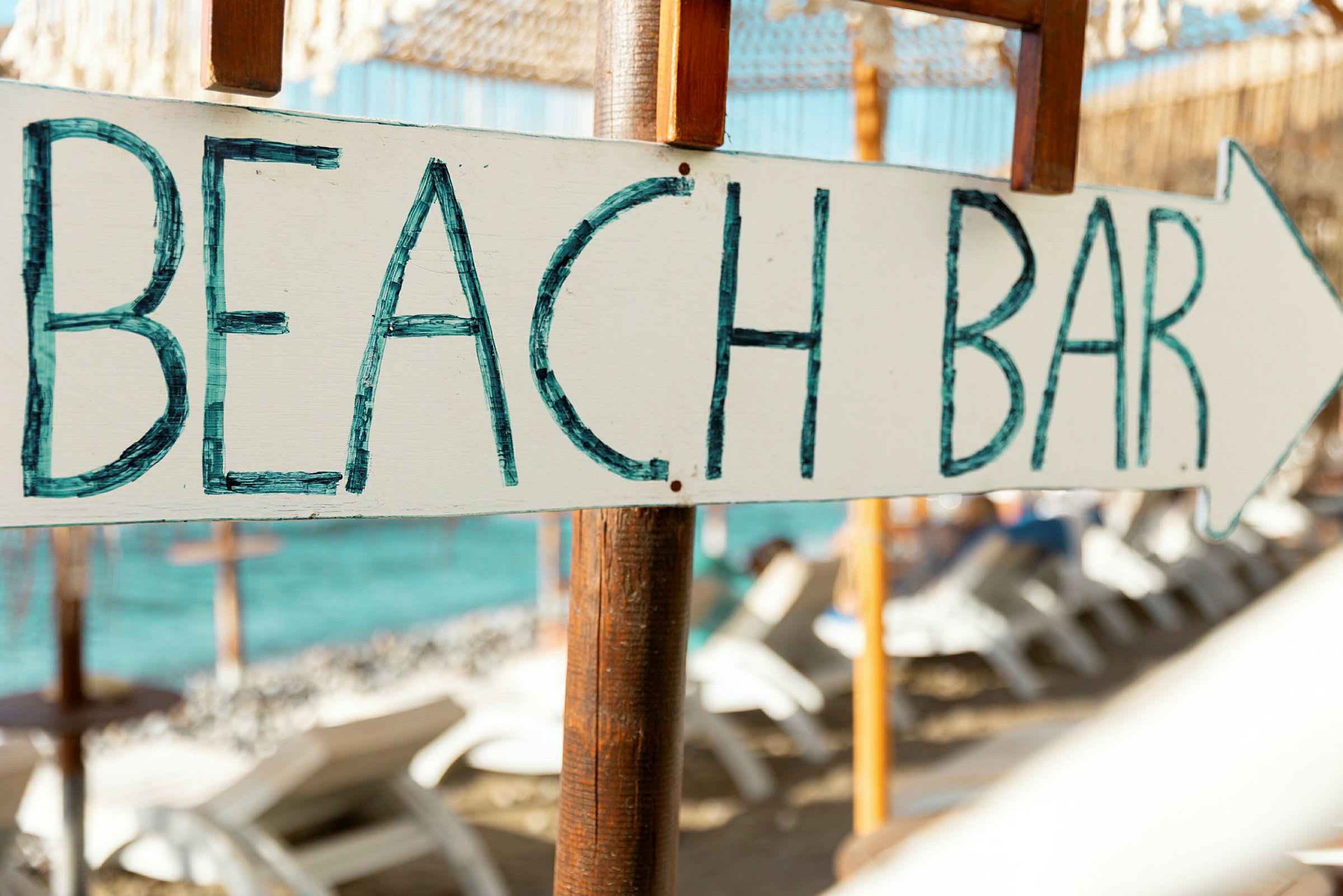 Skilt med teksten 'BEACH BAR' og en pil, i bakgrunnen stranden med solsenger og havet. (AI-generert tekst)