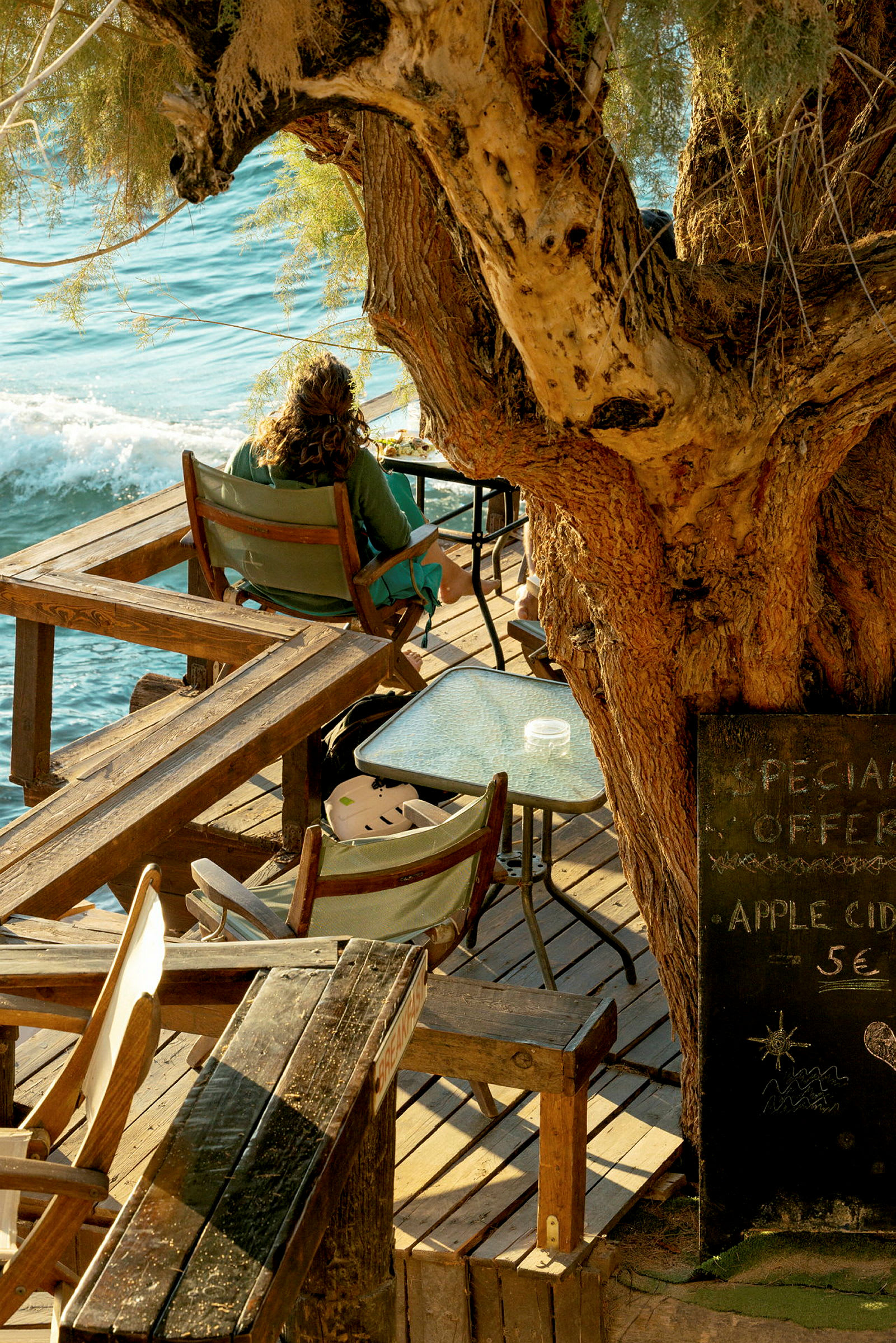 En person sitter på en treterrasse ved havet med et stort tre i nærheten og bord og stoler rundt. (AI-generert tekst)