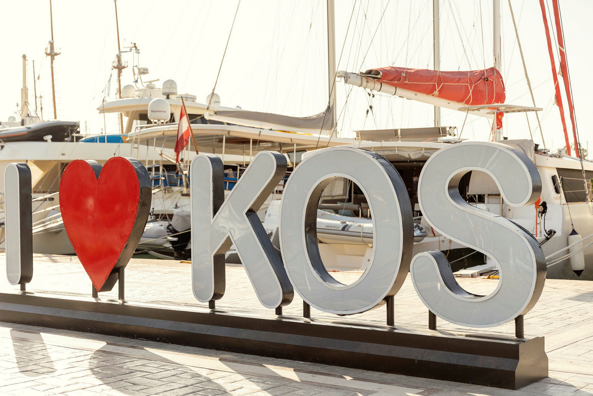 Skilt med teksten 'I ♥ KOS' foran båter i en havn. (AI-generert tekst)