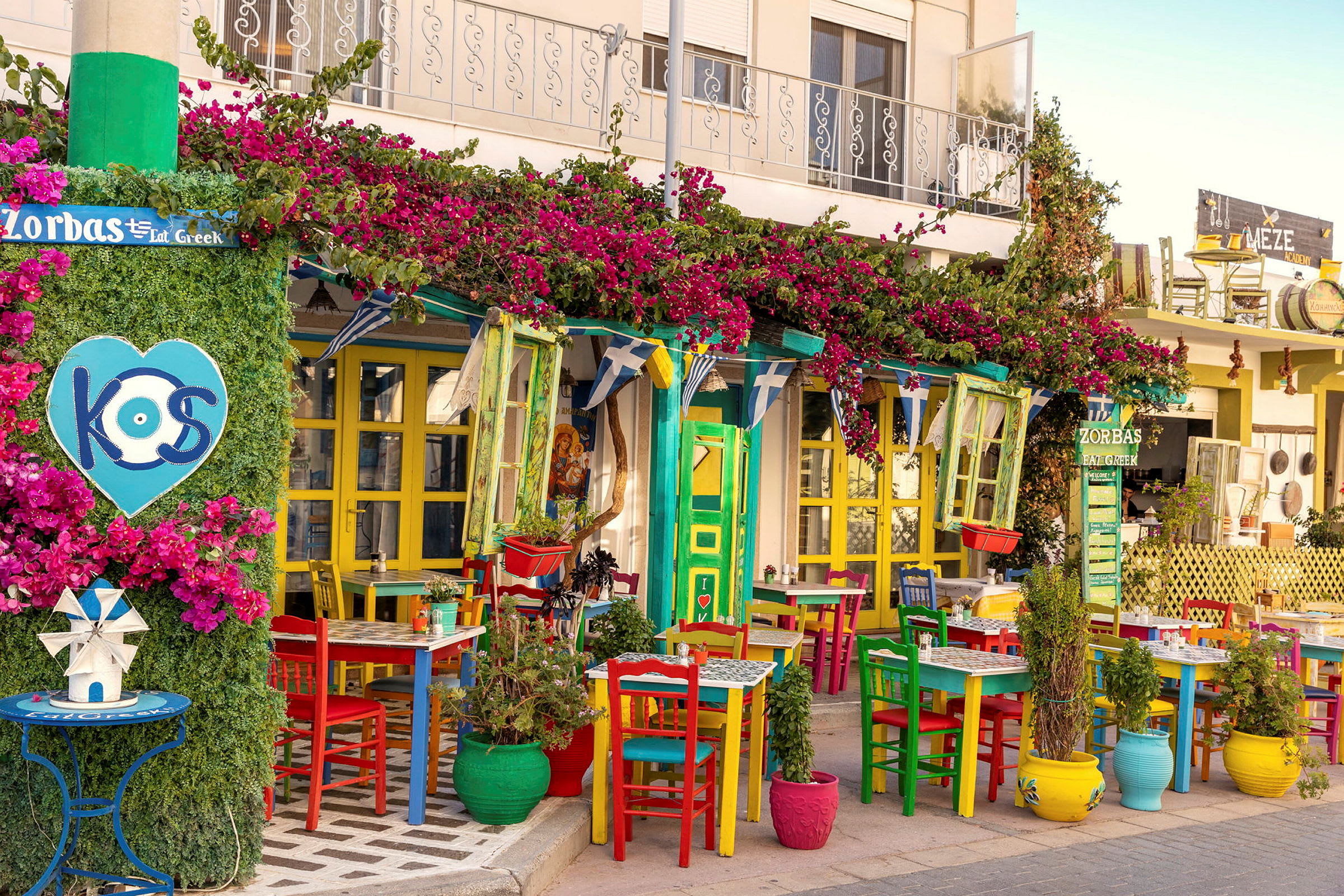 Fargerike stoler og bord utenfor en restaurant dekorert med blomster og vegetasjon. (AI-generert tekst)