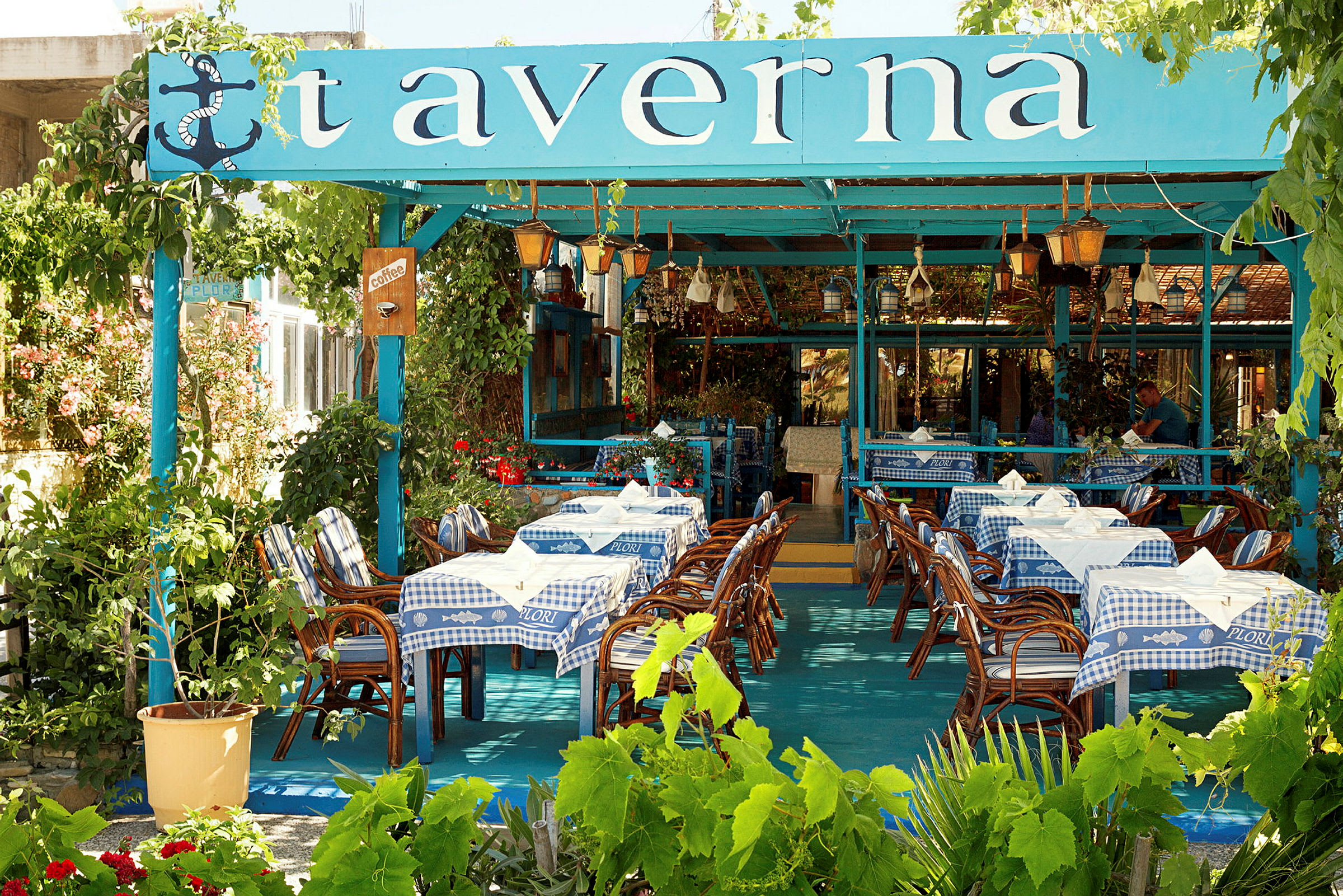 En tradisjonell taverna med blå og hvite duker, bord og stoler i tre, samt vegetasjon rundt. (AI-generert tekst)