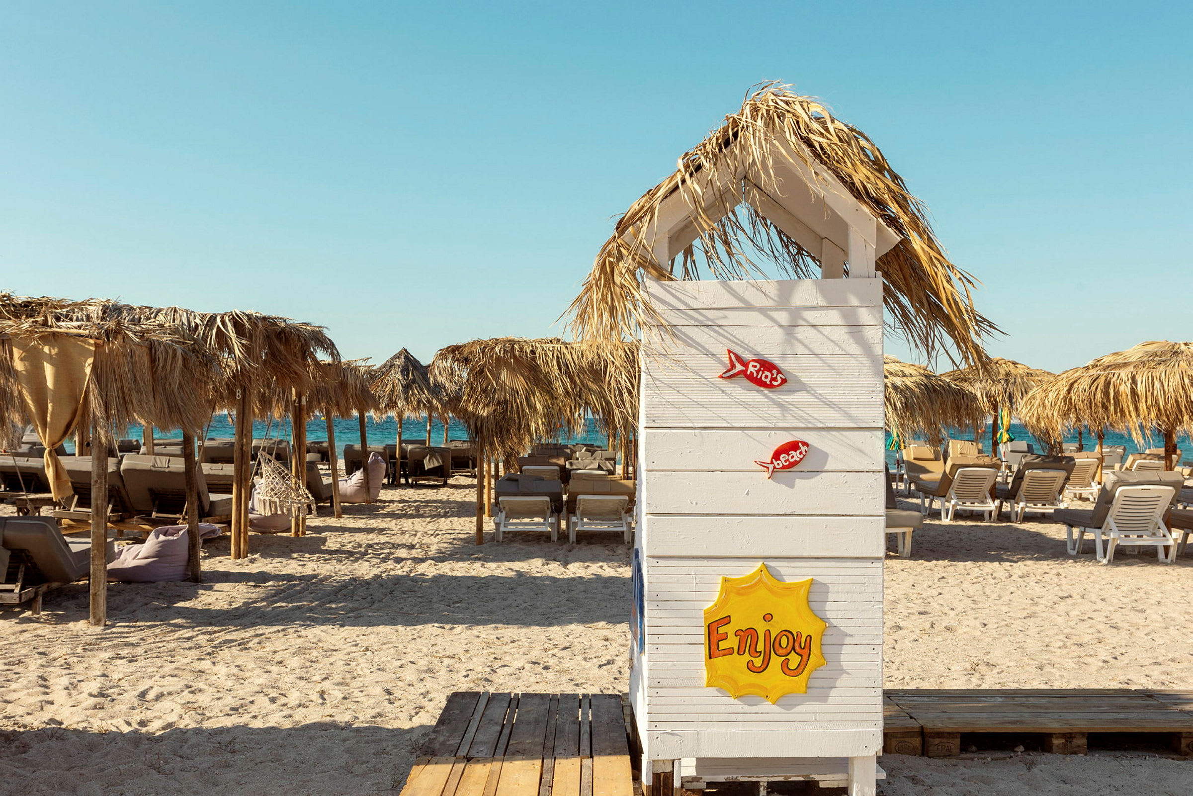 En strand med solsenger og parasoller laget av halm samt en hvit treskjerm nærmest i bildet med teksten «Enjoy». (AI-generert tekst)