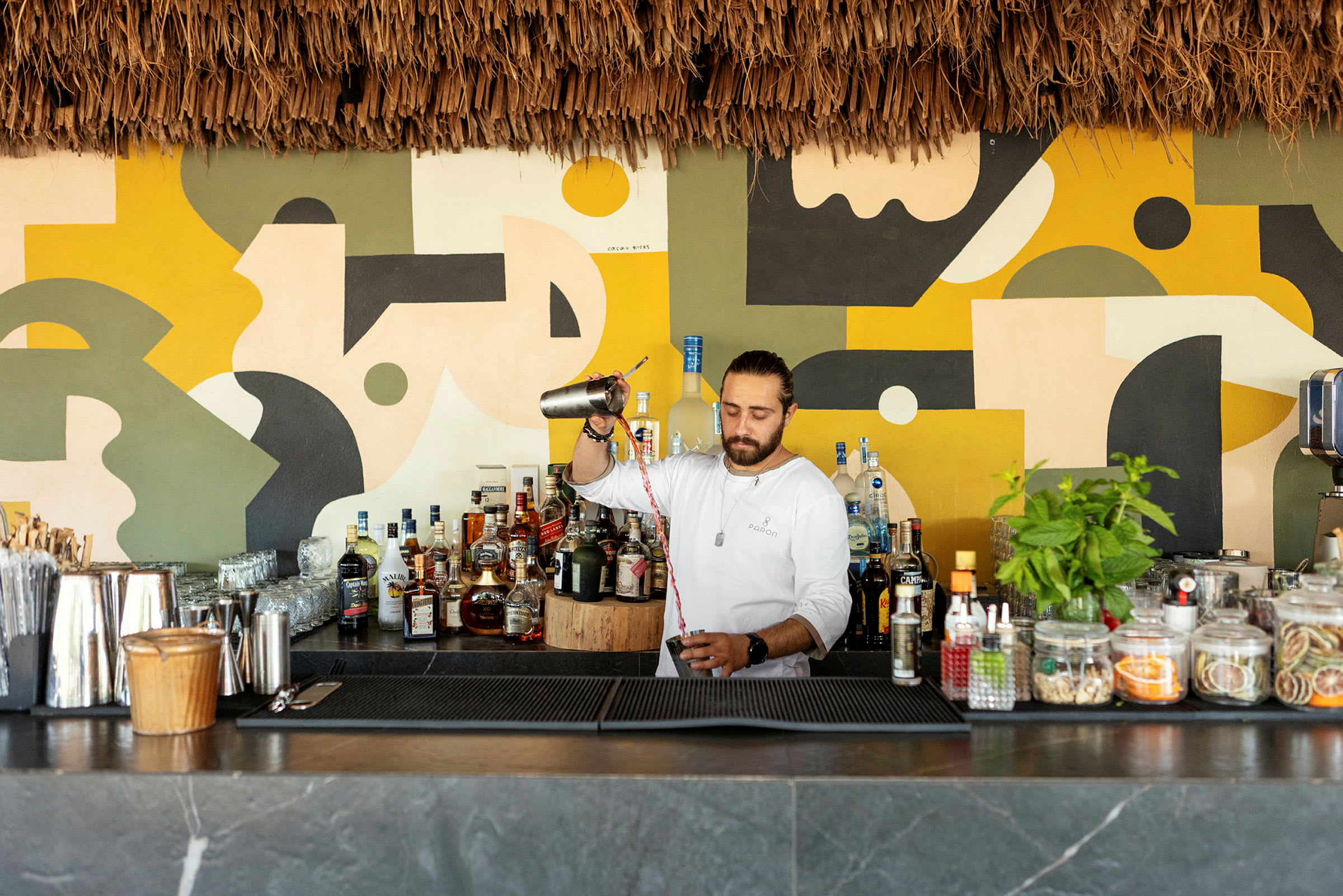 En bartender forbereder en drink bak en bar med fargerike veggdekorasjoner. (AI-generert tekst)
