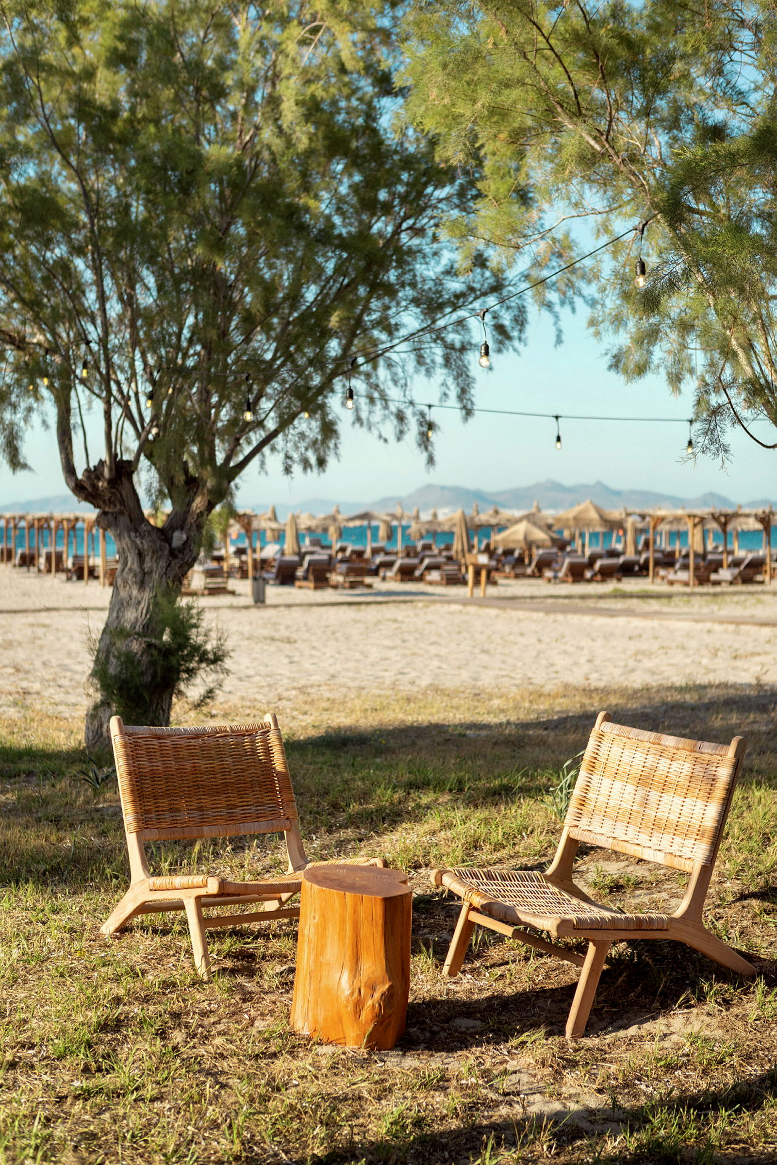 To rottingstoler og et lite bord av tre står på gress nær en strand med parasoller. (AI-generert tekst)