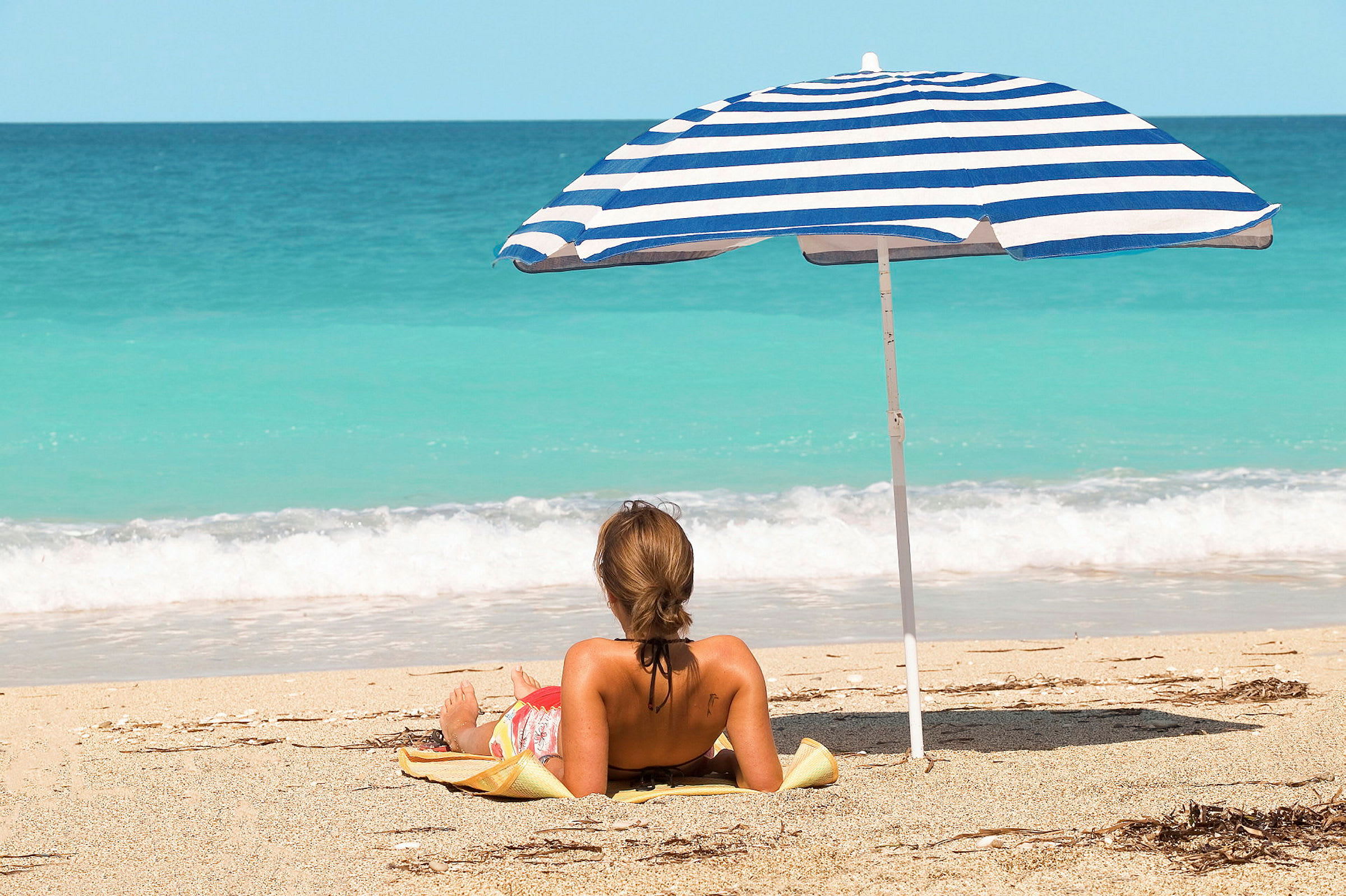 En person ligger på en strand under en blå og hvit parasoll nær havet. (AI-generert tekst)
