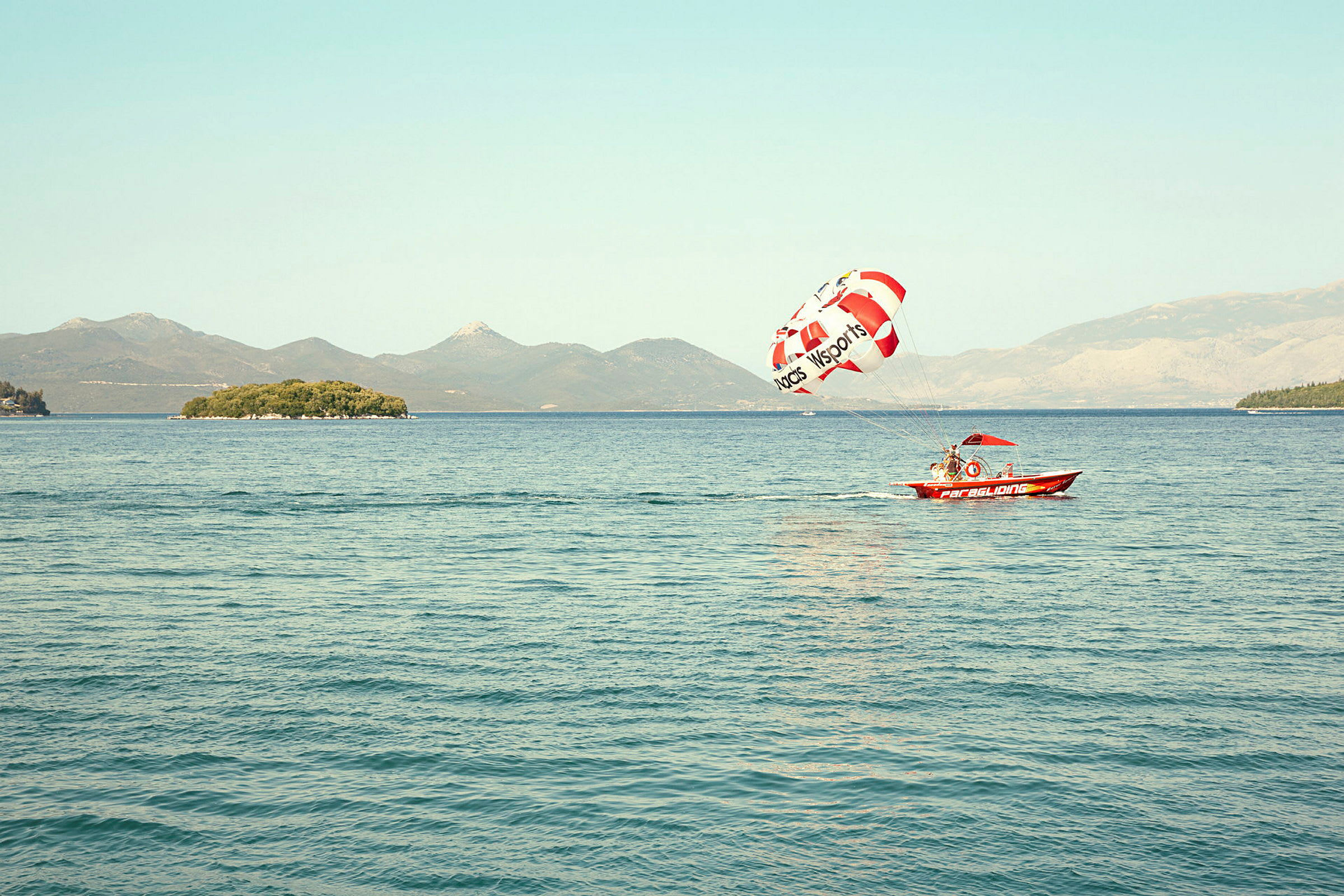 En liten motorbåt trekker en parasailingskjerm over vannet med en øy i bakgrunnen. (AI-generert tekst)