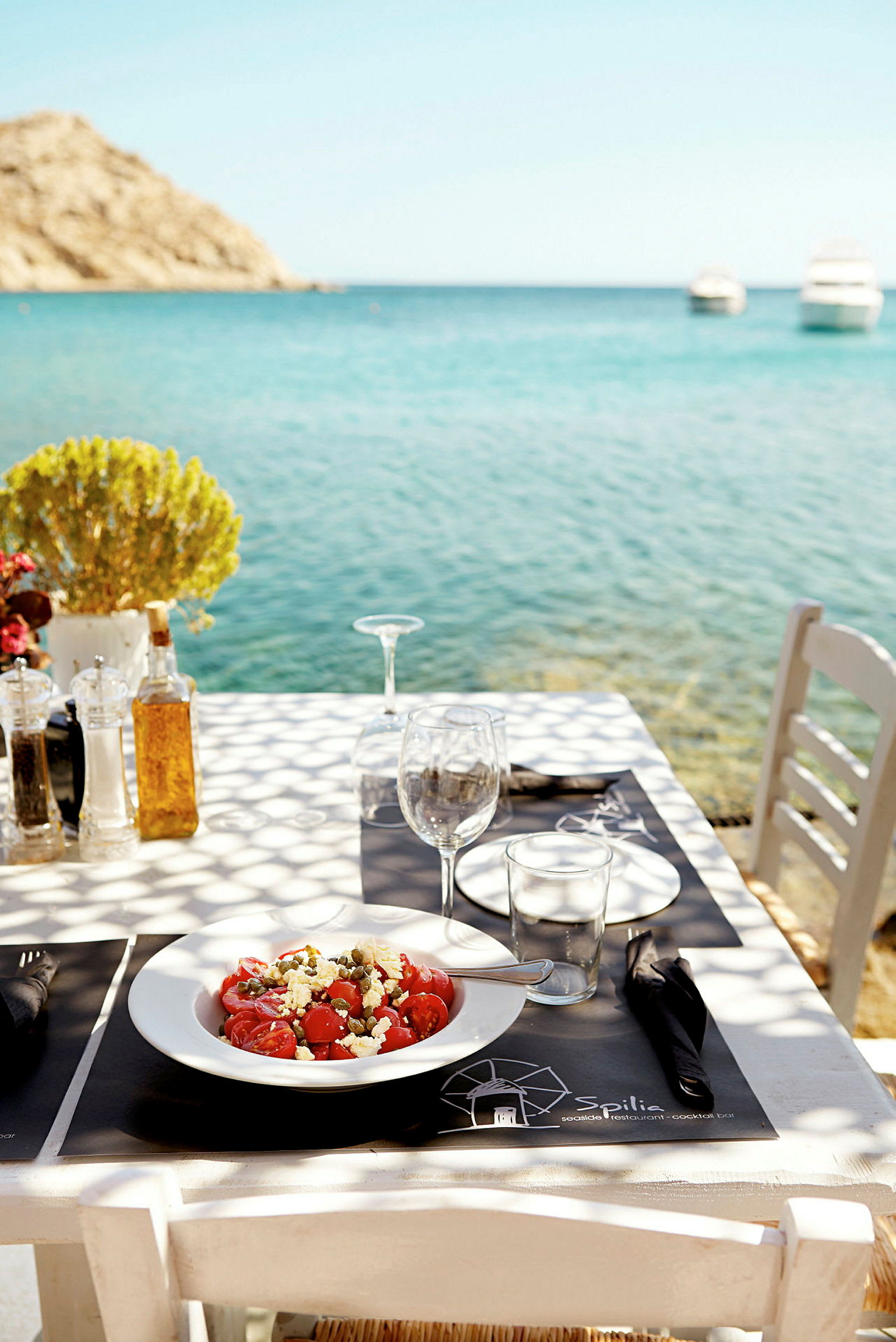 Et spisebord ved havet med en tallerken salat og vinflasker samt glass. Bakgrunnen viser blått hav og båter. (AI-generert tekst)