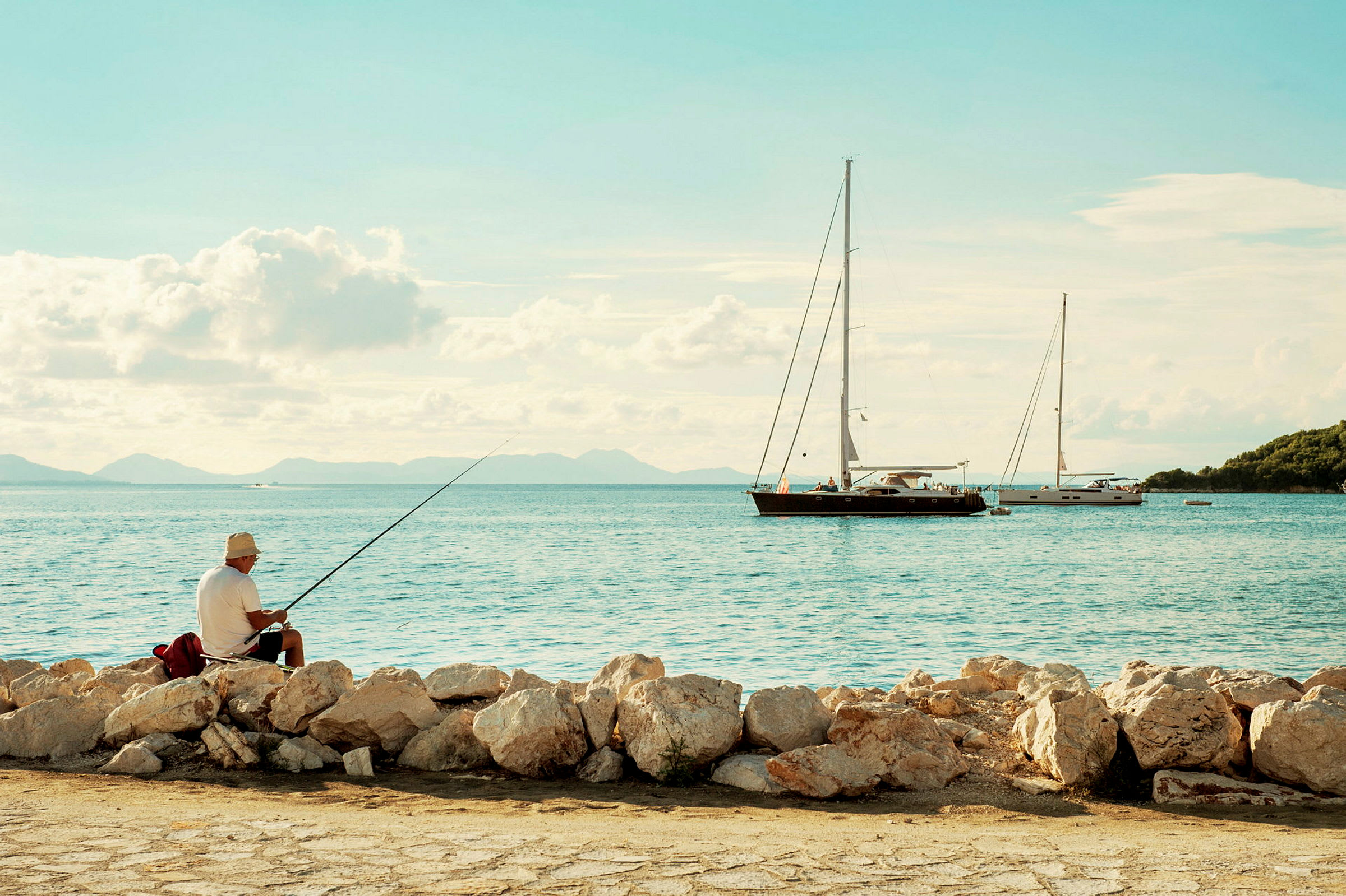 En person sitter og fisker ved en steinete strand, med seilbåter i bakgrunnen. (AI-generert tekst)
