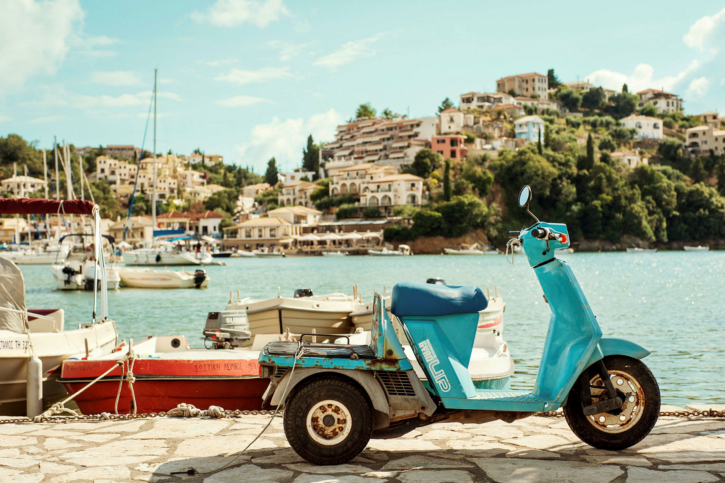 En blå scooter står parkert ved en havn med båter og en fjellskråning i bakgrunnen. (AI-generert tekst)