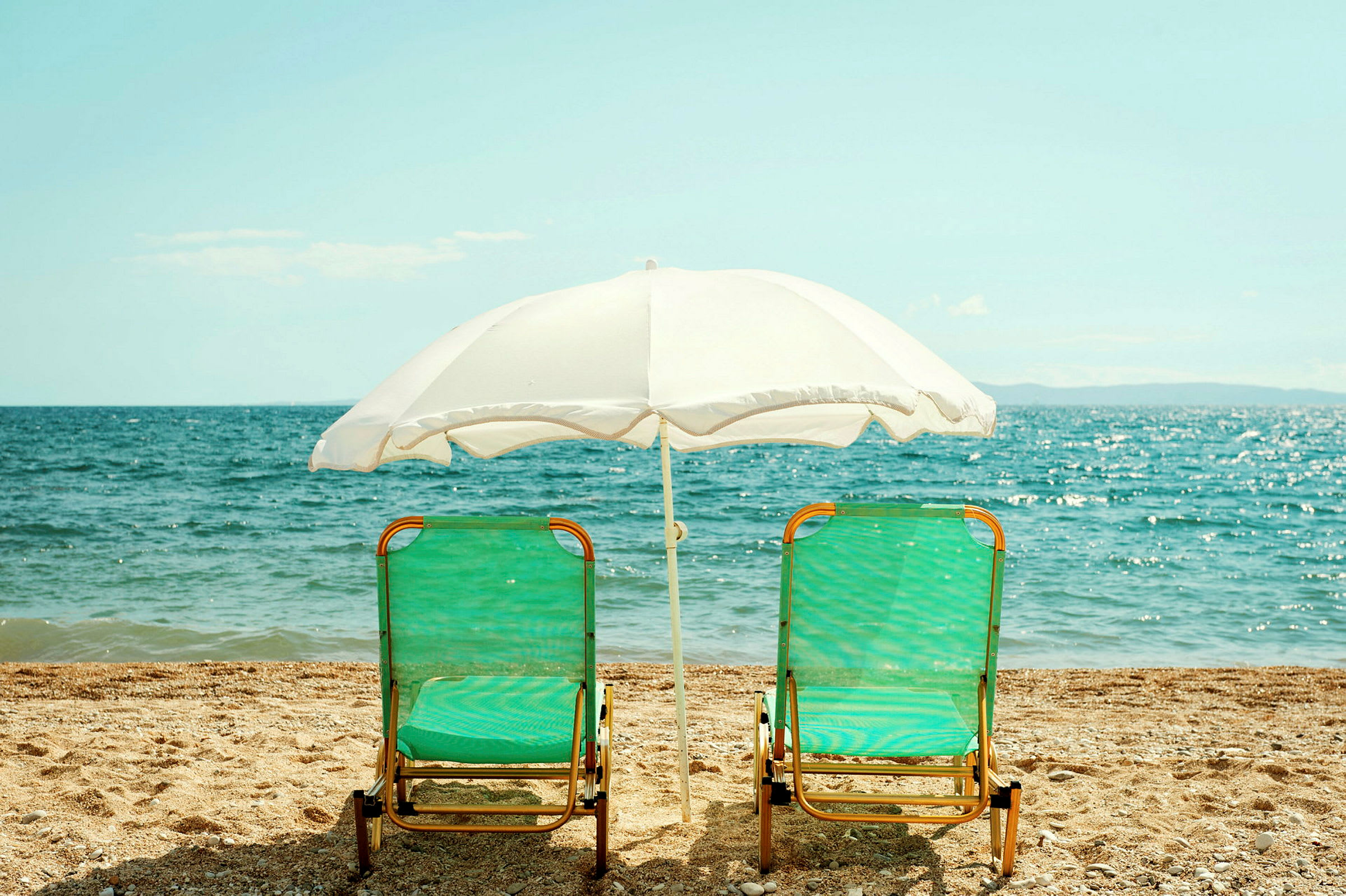To solsenger med grønne sitteputer og en hvit parasoll står på stranden foran sjøen. (AI-generert tekst)
