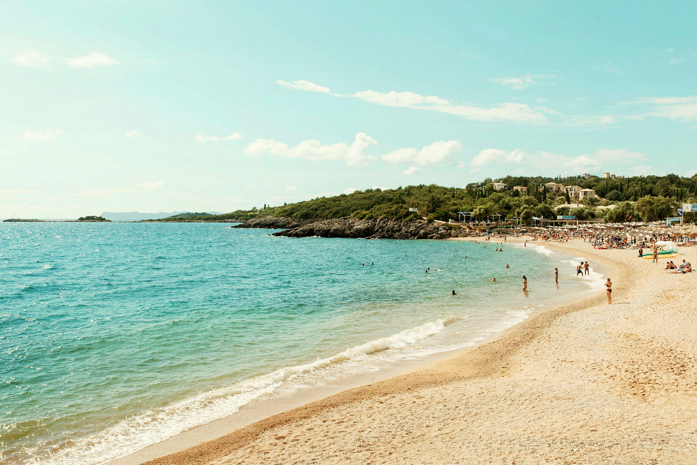 Strand med sand og klart blått vann, omgitt av grøntområder og folk som bader. (AI-generert tekst)