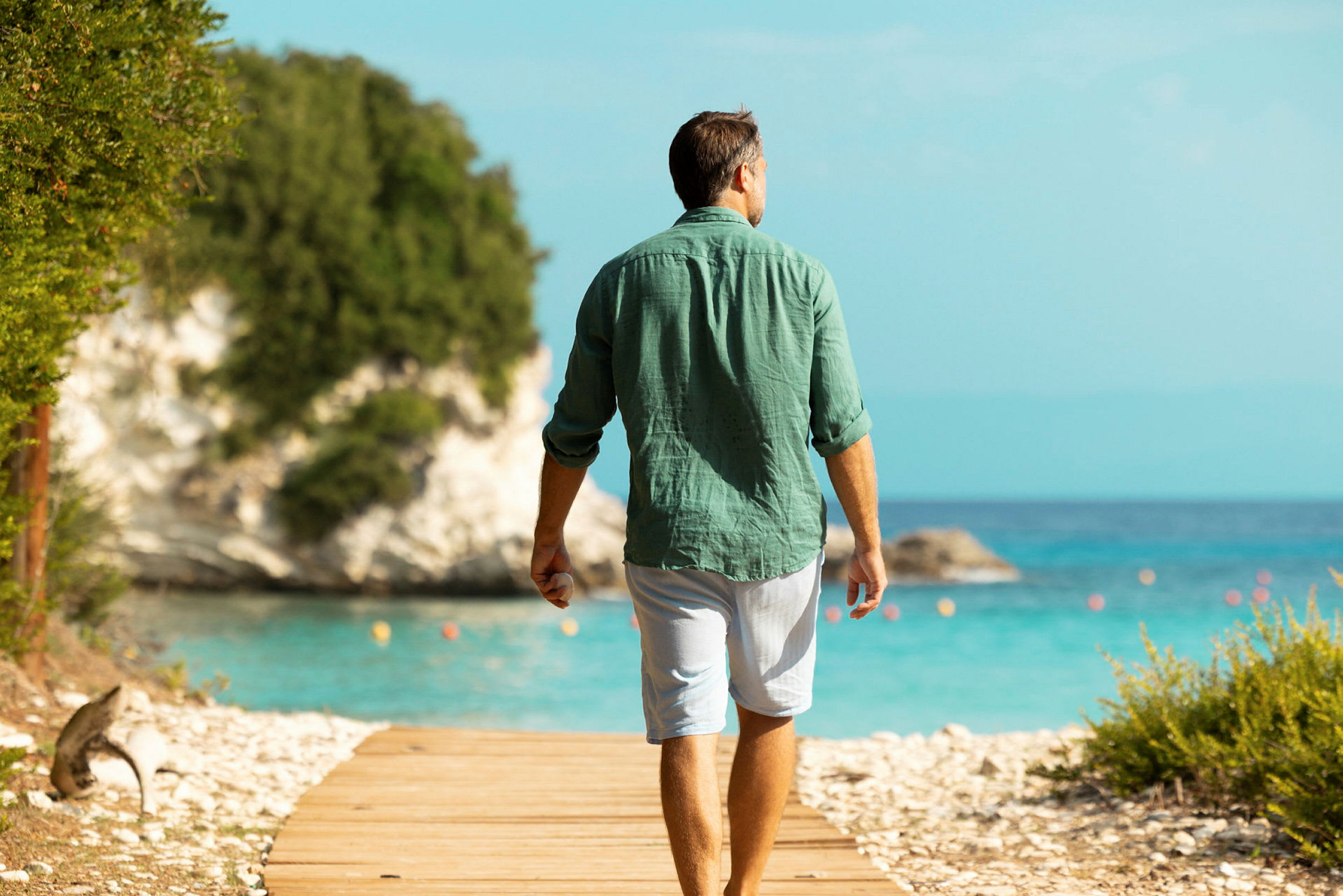 En person går på en tresti ved siden av havet med frodige klipper i bakgrunnen. (AI-generert tekst)