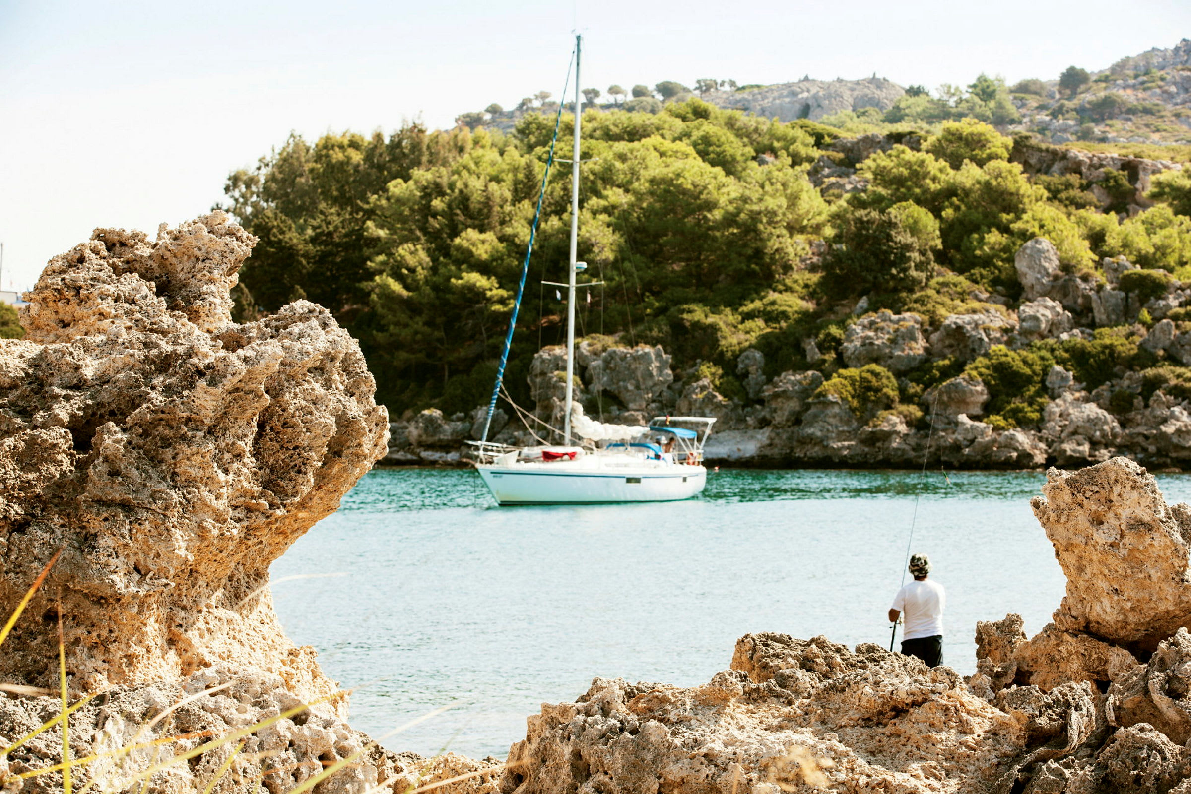 En seilbåt ligger fortøyd ved en fredelig vik omgitt av klipper og skog, med en person som fisker fra stranden. (AI-generert tekst)