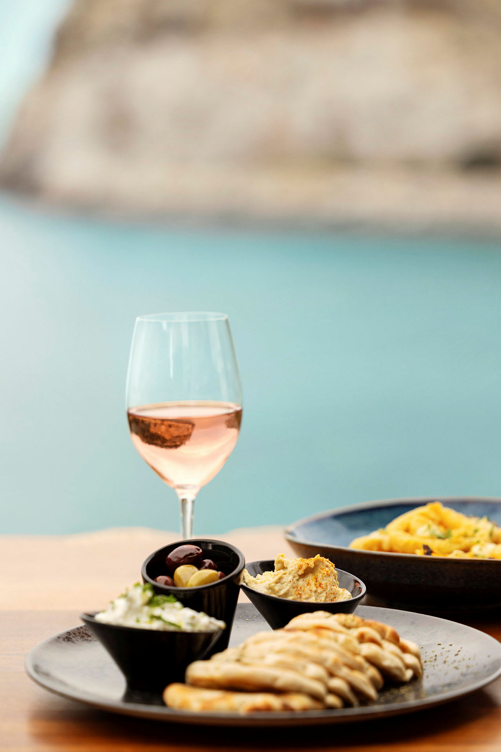 Et glass rosévin ved siden av en tallerken med brødskiver, en olivenskål og kremete pålegg. (AI-generert tekst)