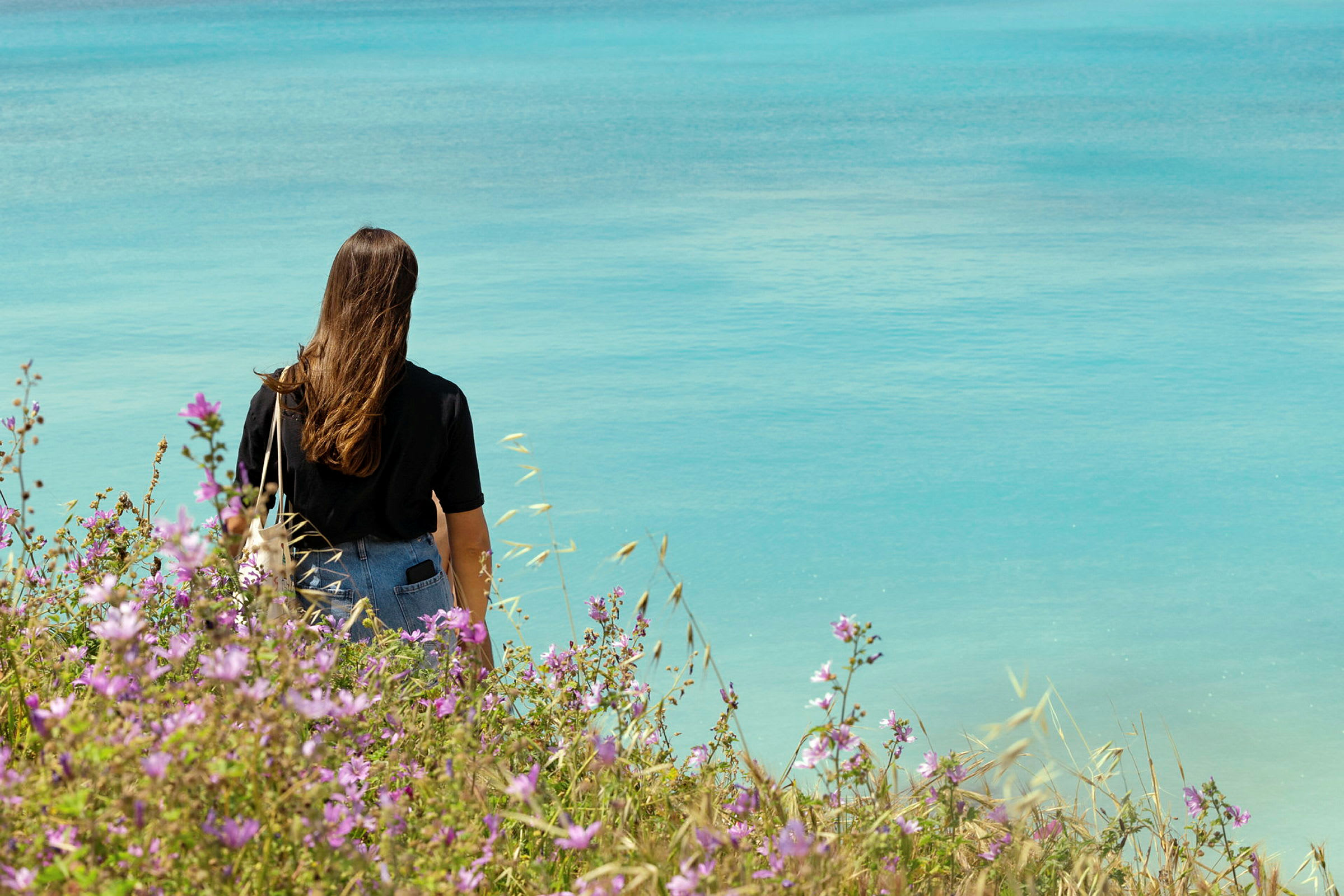 En kvinne står ved kysten med ryggen til kameraet og ser mot havet, omgitt av blomstrende planter. (AI-generert tekst)
