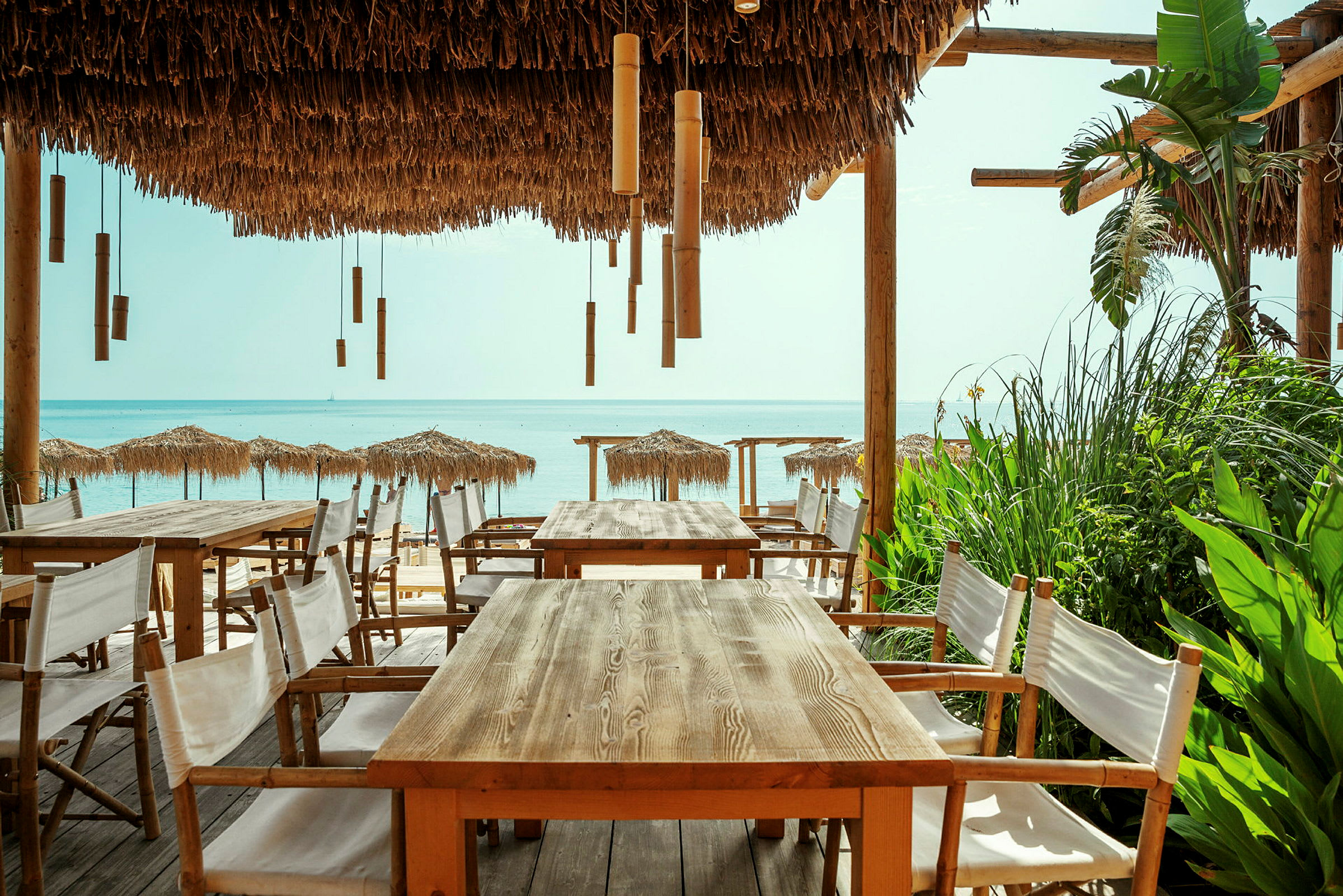 Utendørs serveringsområde med trebord og stoler under en pergola med stråtak, med utsikt over stranden og havet. (AI-generert tekst)