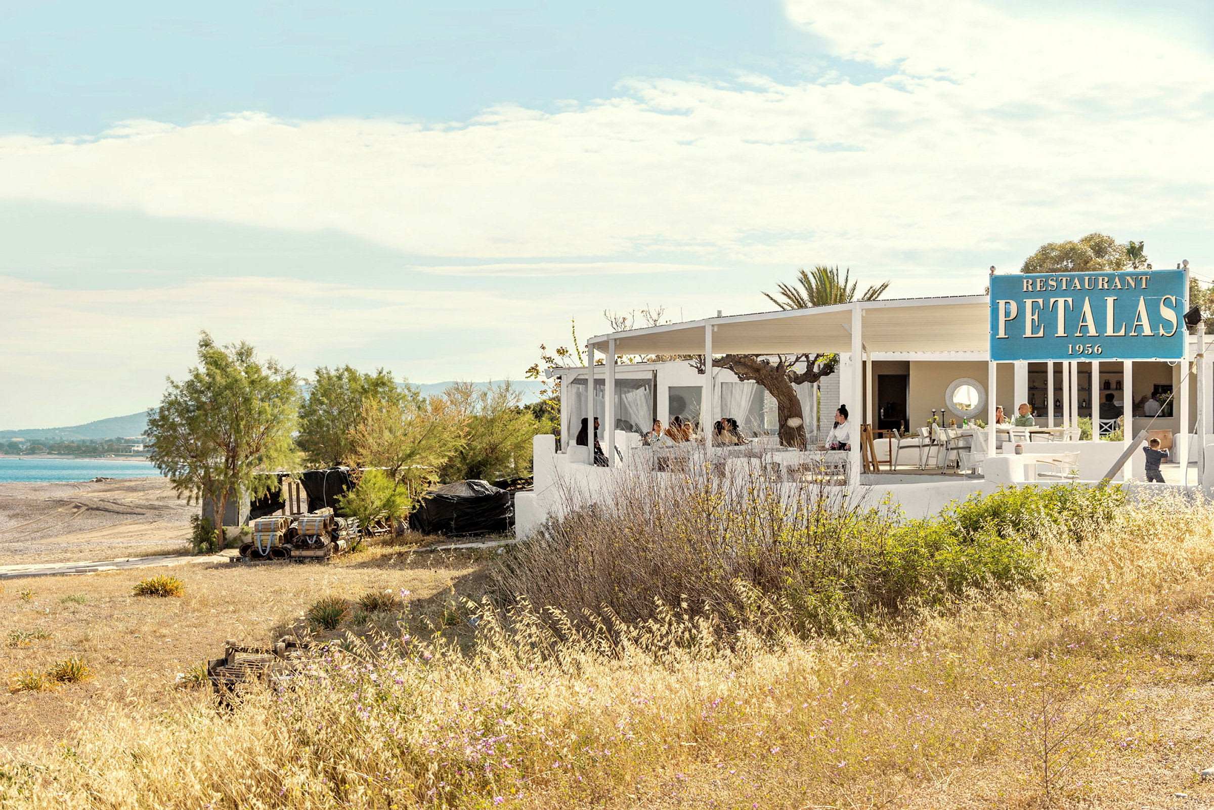 En restaurant med hvit bygning nær en strand med utendørs servering under tak. (AI-generert tekst)
