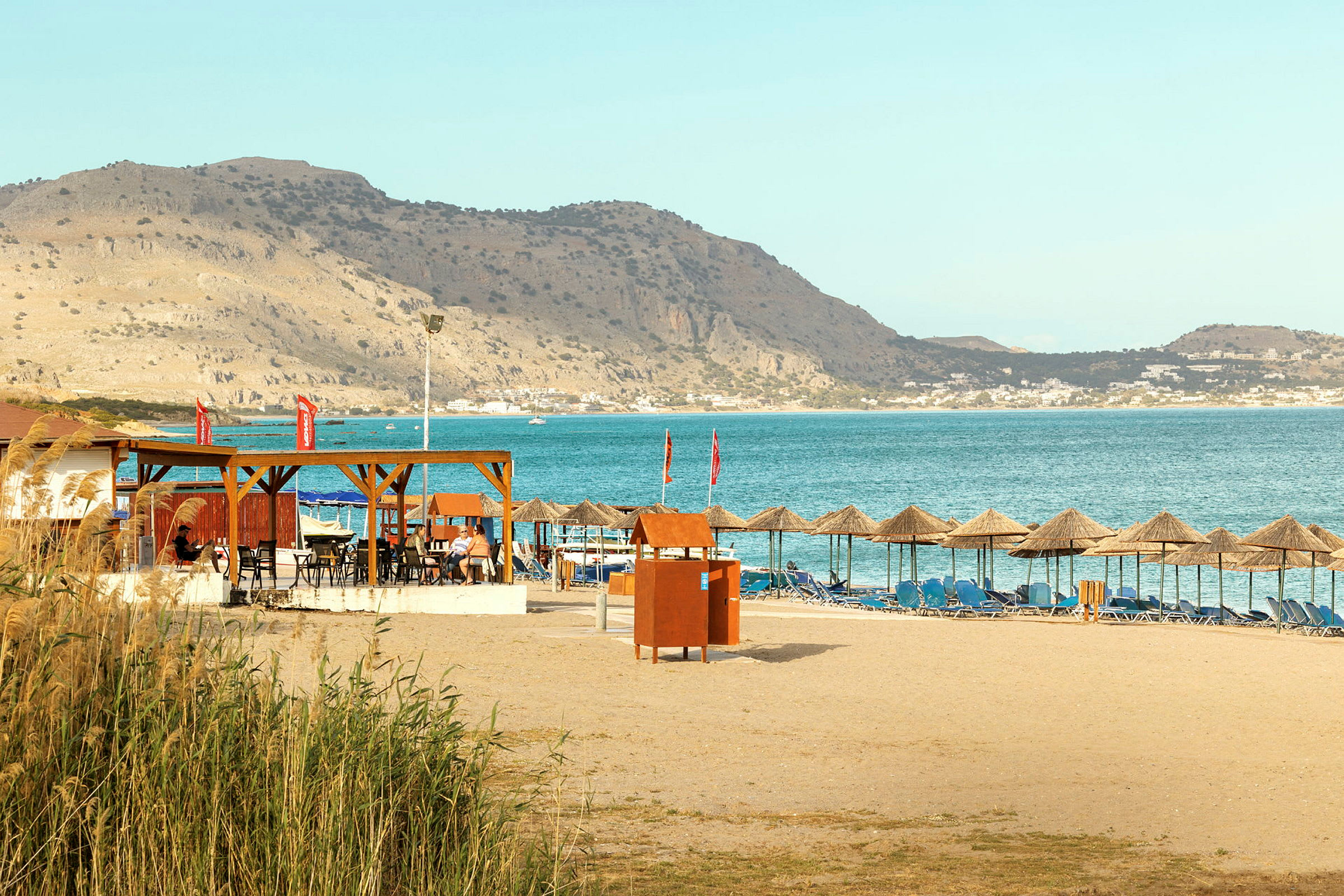 Sandstrand med solsenger og parasoller, samt en restaurant med utsikt over havet og fjellene i bakgrunnen. (AI-generert tekst)