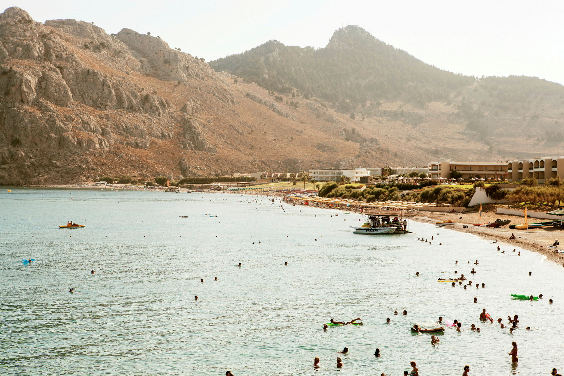 En strand ved en bukt omgitt av fjell, med mennesker som svømmer i vannet og soler seg på stranden. (AI-generert tekst)