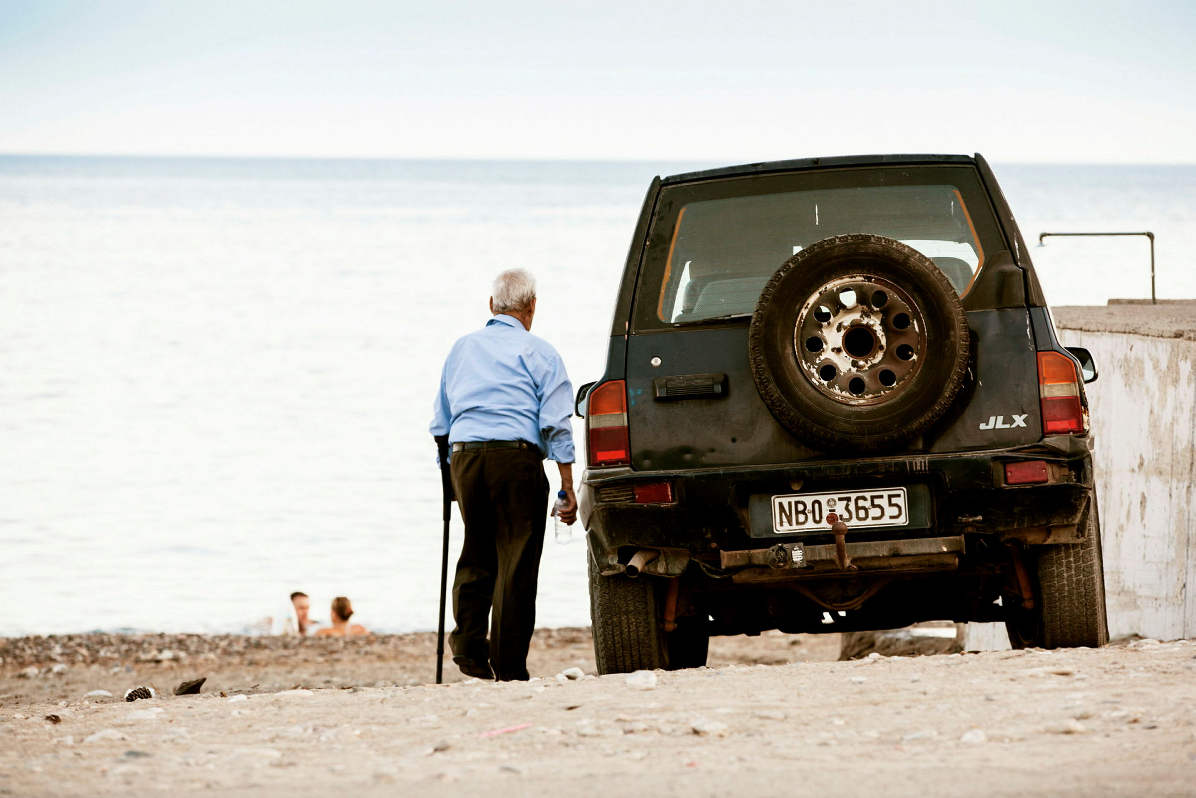 En eldre person står på en strand ved siden av en svart bil med reservedekk på baksiden. (AI-generert tekst)