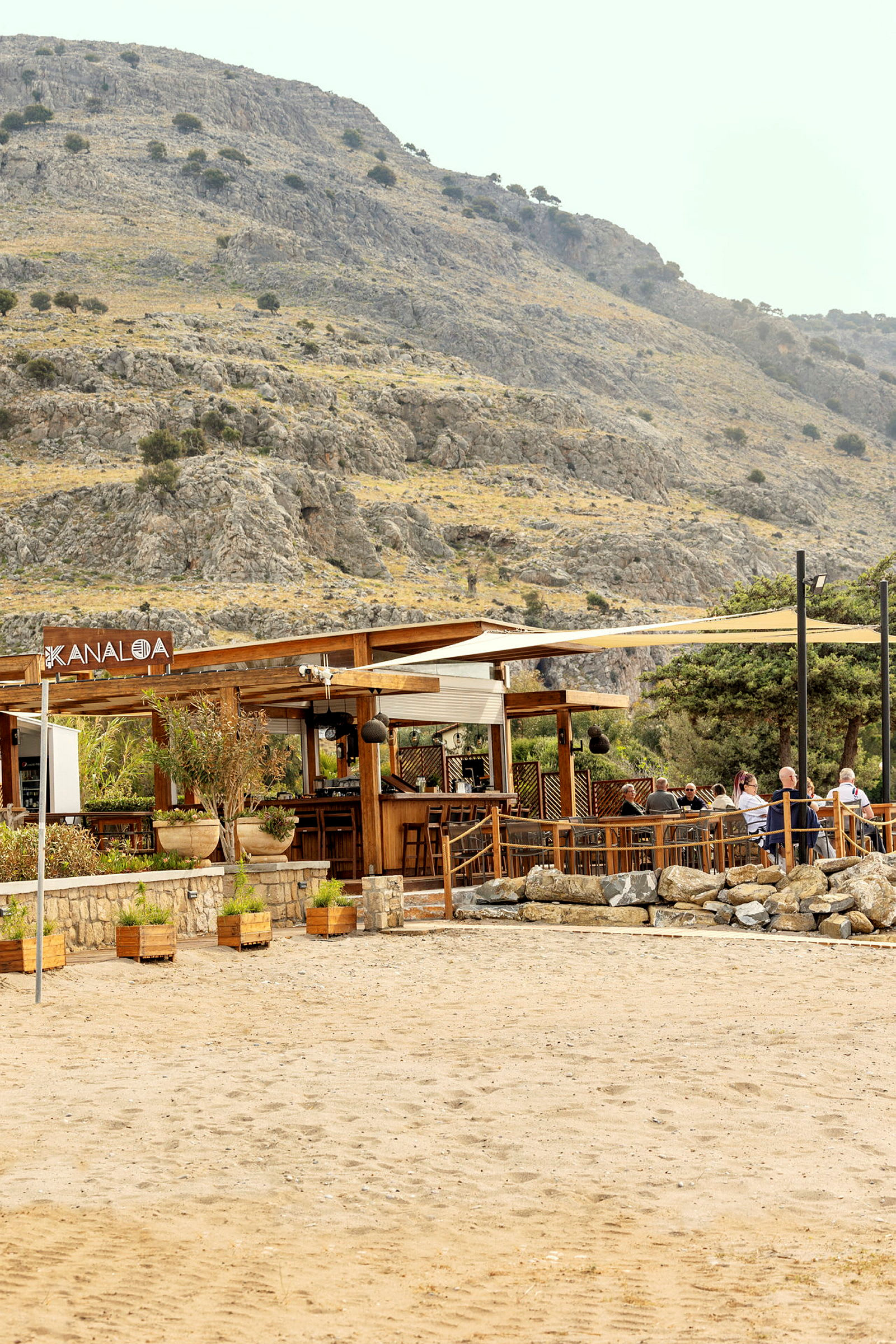 En strandbar med tremøbler og parasoller, omgitt av en fjellrik setting. (AI-generert tekst)